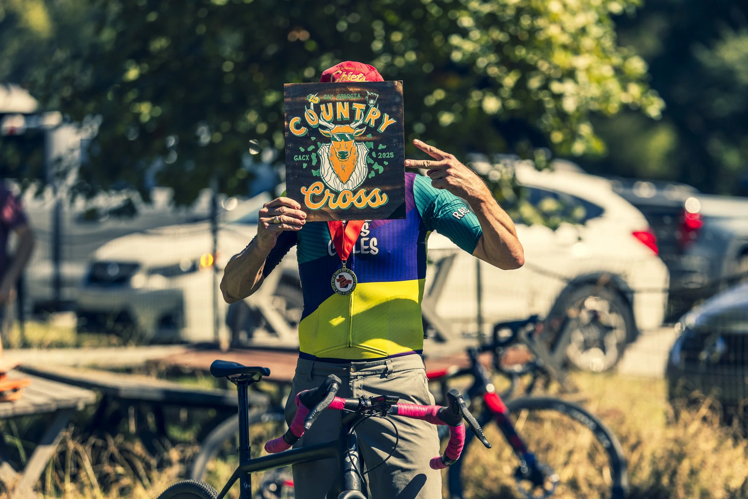 Cyclist holding a sign that says "Country Cross" with a graphic of a goat's head, wearing a yellow and blue multicultural cycling jersey, standing beside a black bicycle with pink handlebar tape at an outdoor event with trees and parked cars in the b