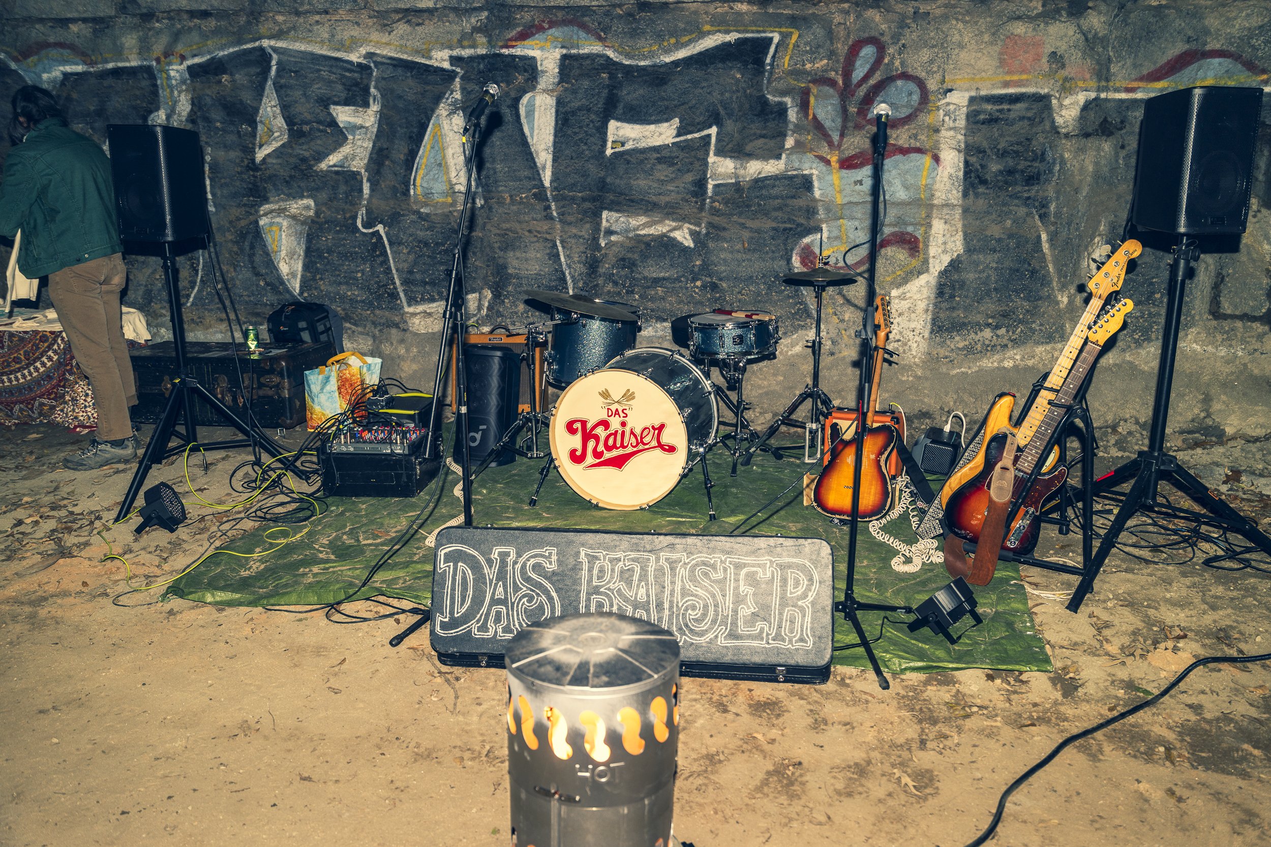 Empty stage set for a band with various guitars, drums, microphones, and speakers, in front of a graffiti-covered concrete wall, outdoors at night, with a lit fire pit in the foreground.