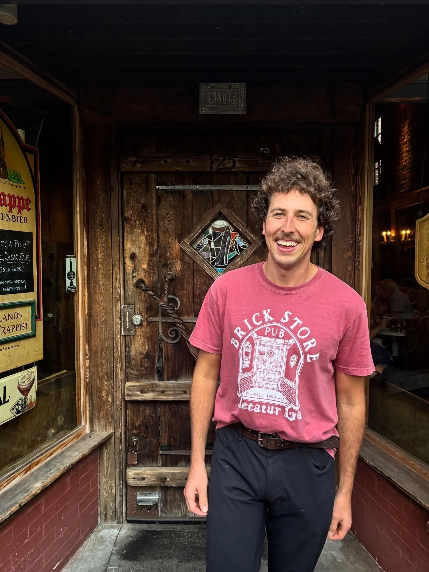 A young man with curly hair and a wide smile standing outside a rustic wooden door at a pub, wearing a pink T-shirt and black pants.