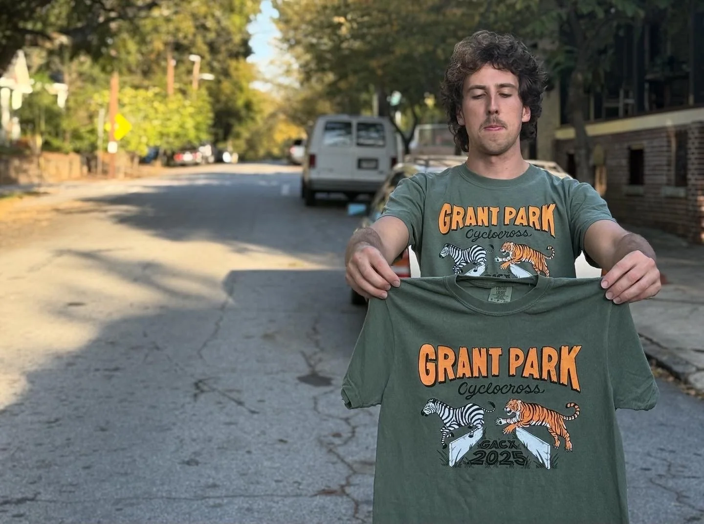 A man standing outdoors on a street holding a green T-shirt with a graphic design and text. The T-shirt features black and white and orange tigers, the words "GRANT PARK," "Cyclocross," and the year "2025." The man is wearing a matching T-shirt with 