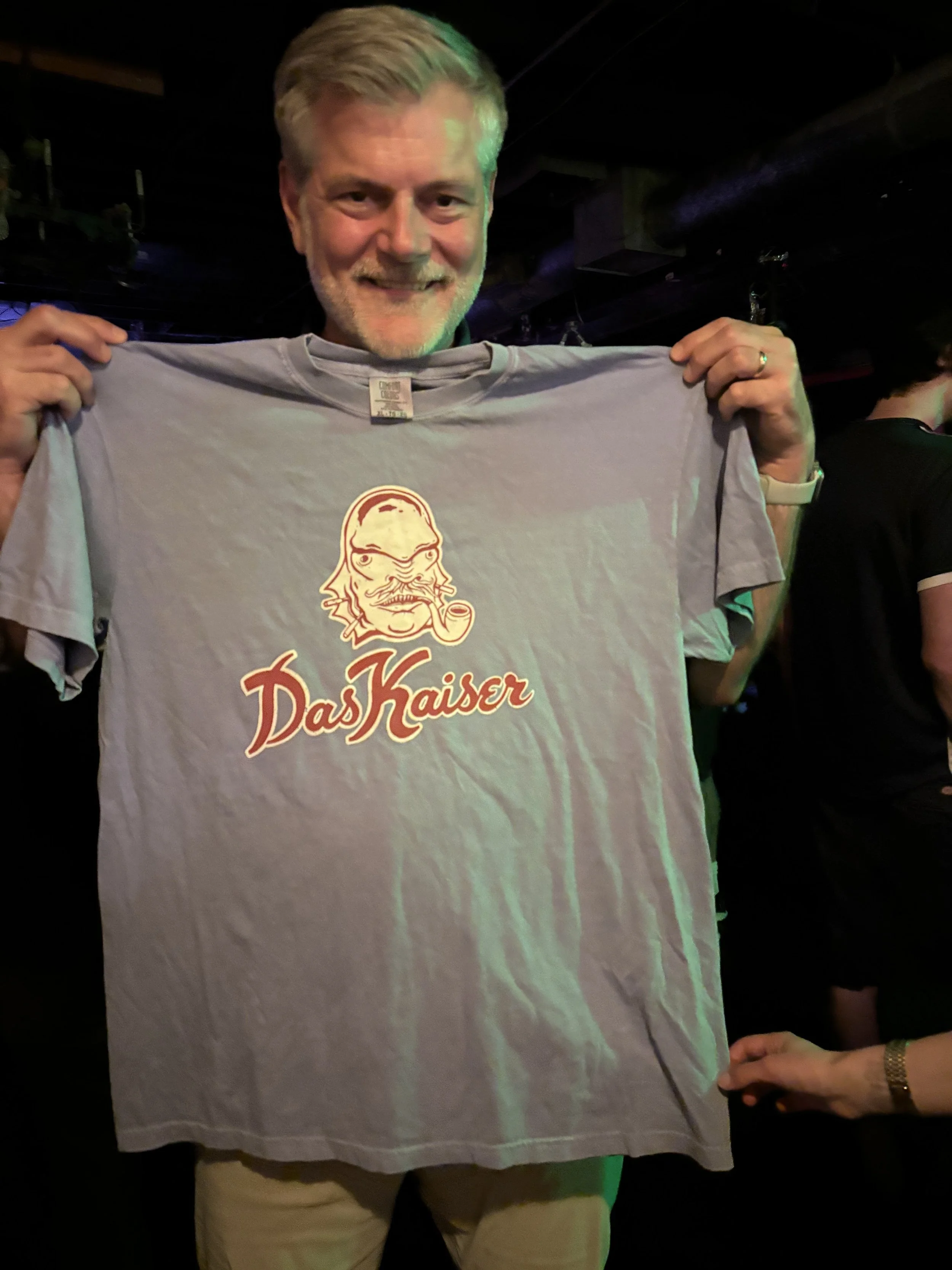 A smiling man holding a gray T-shirt with a caricature of a bearded man smoking a pipe and the words 'Das Kaiser' printed on it.