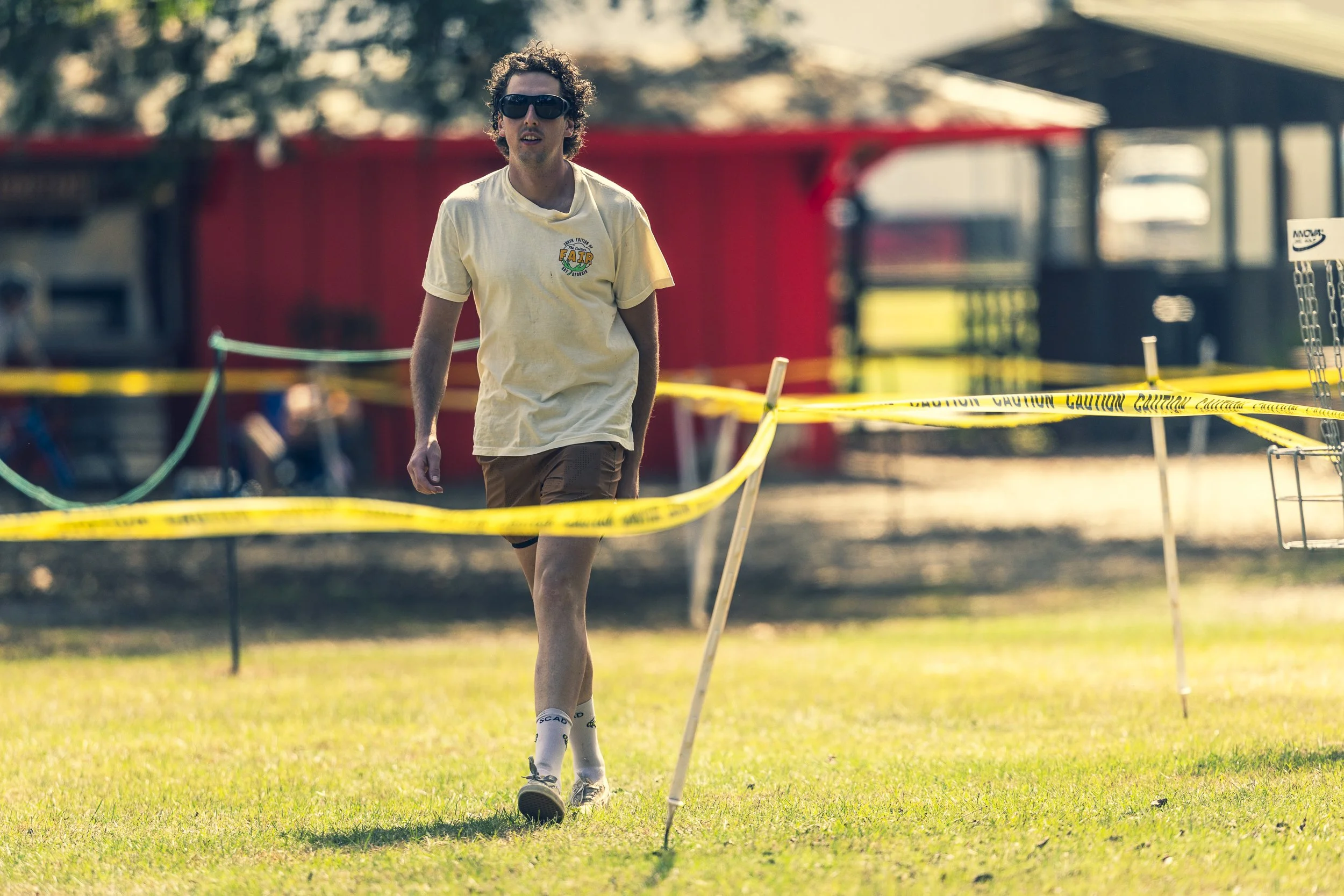 A young man with curly hair wearing sunglasses, a yellow T-shirt, and shorts walking through a field sectioned off with yellow caution tape.
