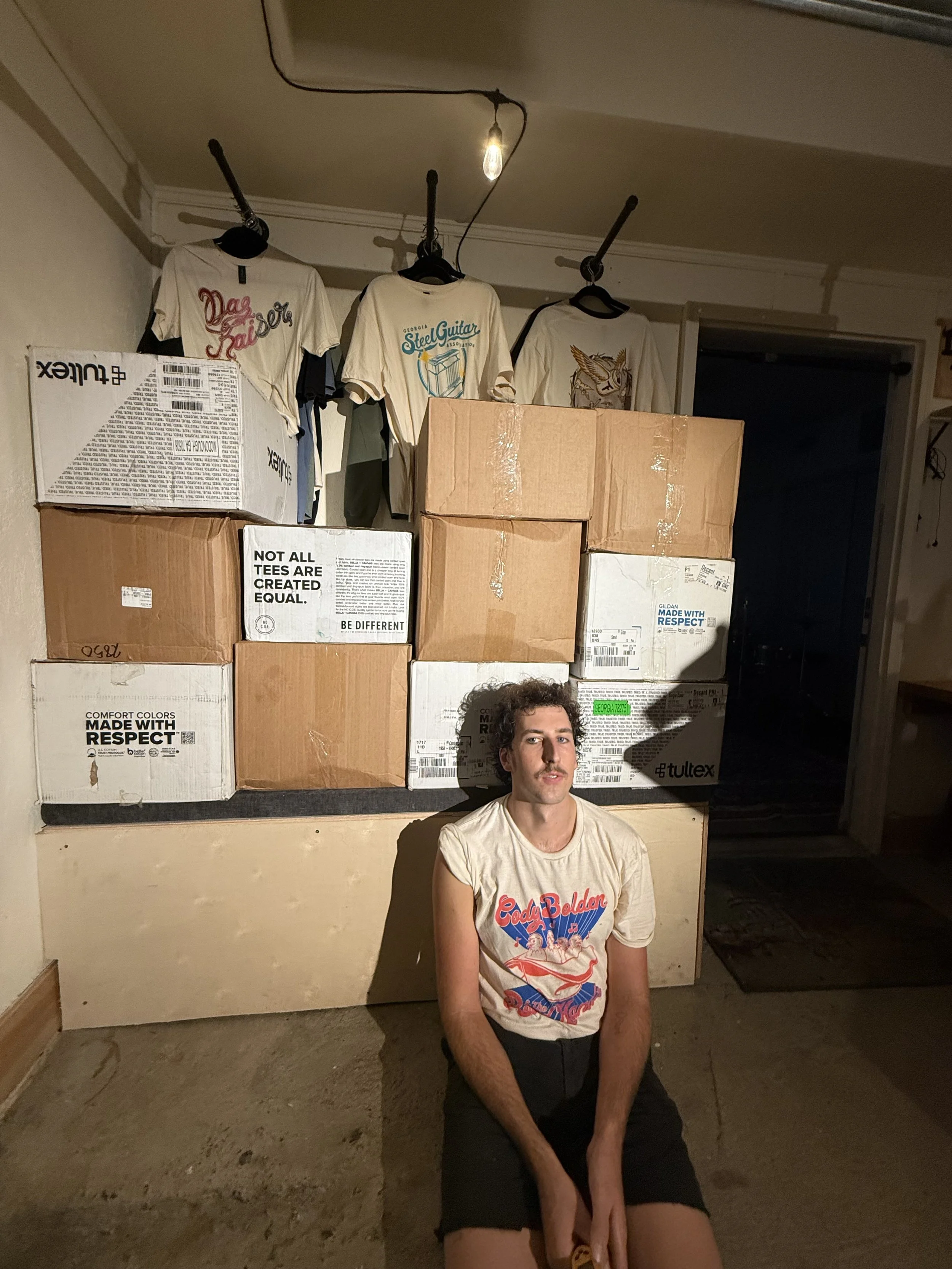 A man sitting on the floor in front of stacked cardboard boxes, with three T-shirts hanging above on the wall, in a room with a hanging light bulb.