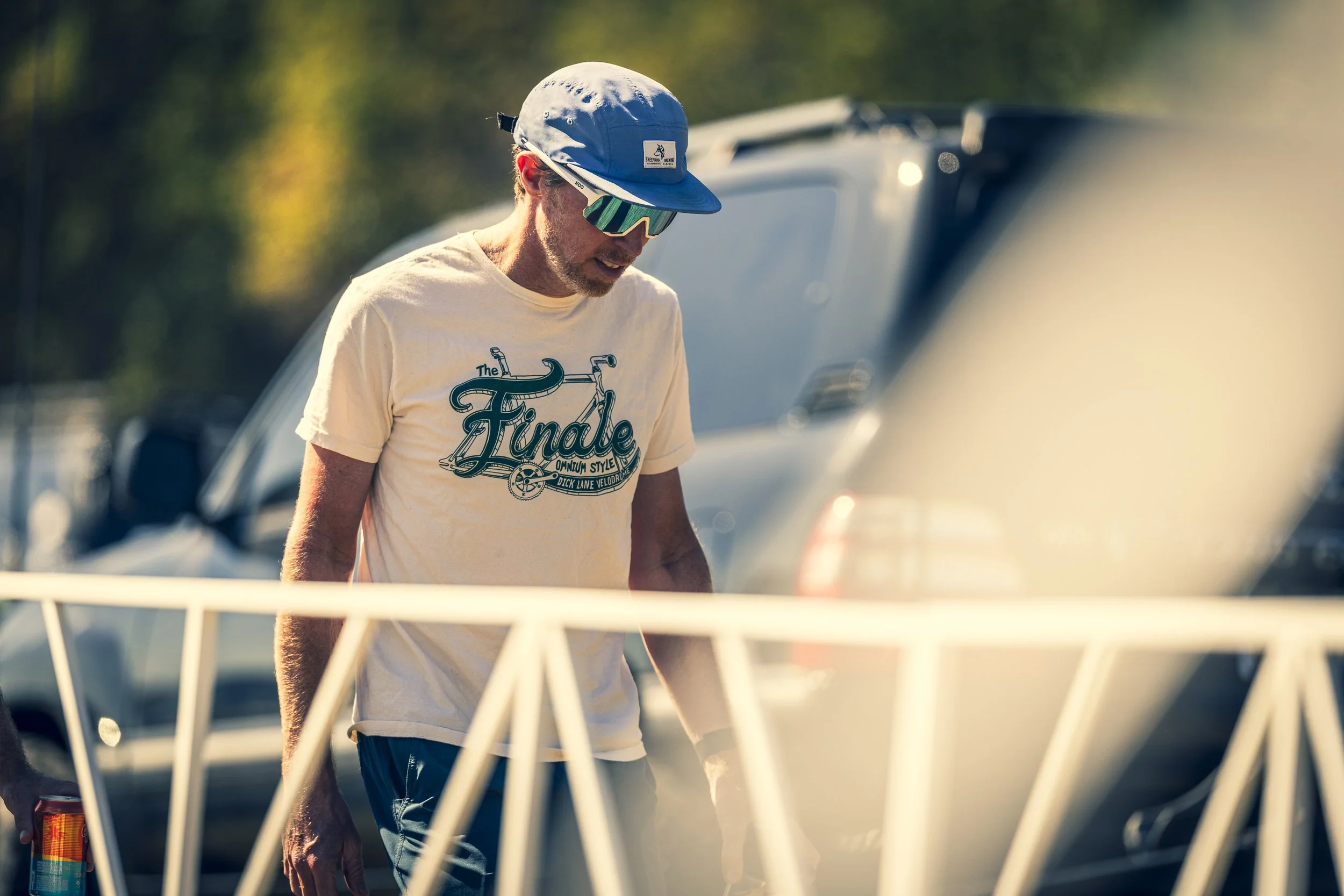 A man wearing a blue cap, reflective sunglasses, and a yellow T-shirt with the word 'The Finale' in graphic print, standing outdoors near parked cars.