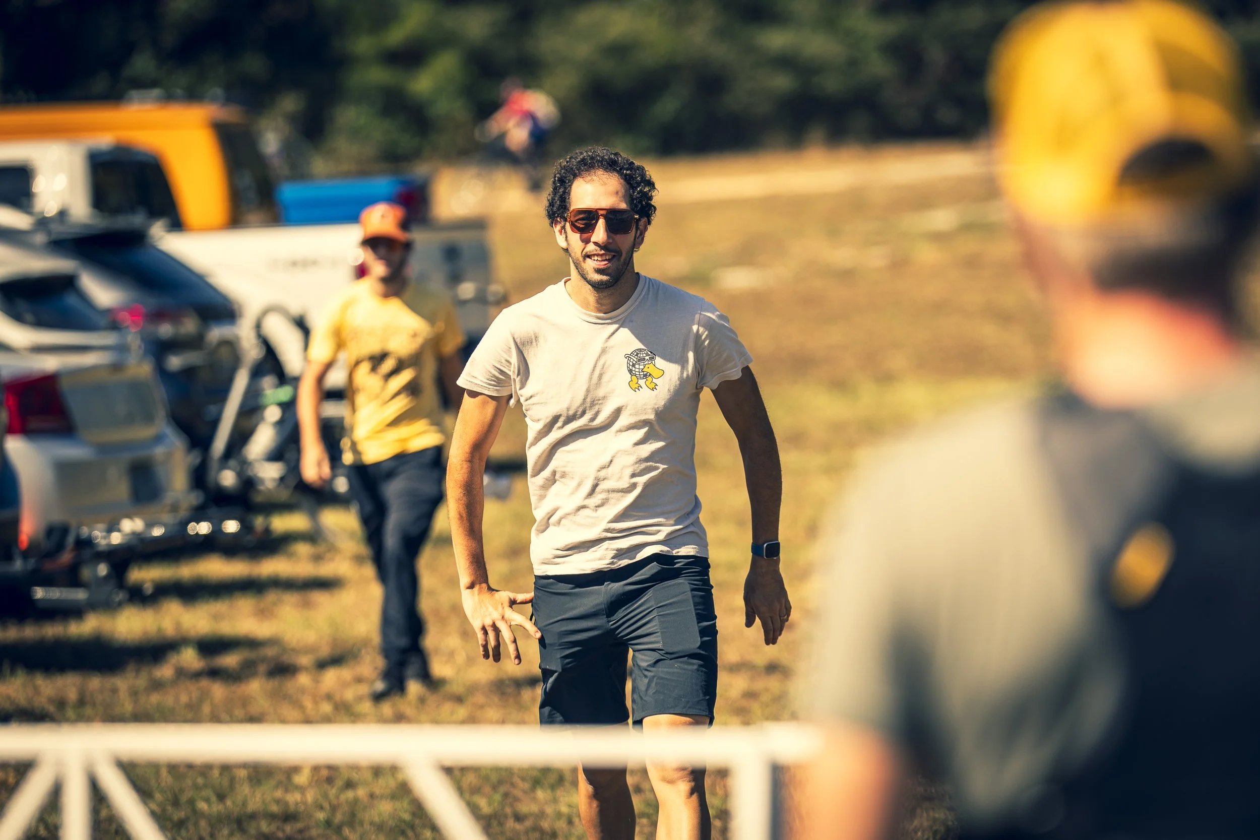 A man with curly hair and sunglasses walking outdoors on a sunny day, with vehicles and other people in the background.