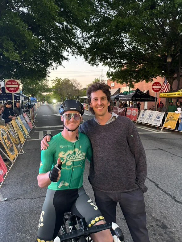 Two men standing together at a street fair, one dressed as a cyclist in a green racing outfit and helmet, giving a thumbs-up, the other in casual clothes with his arm around the cyclist, both smiling, with booths and trees in the background.