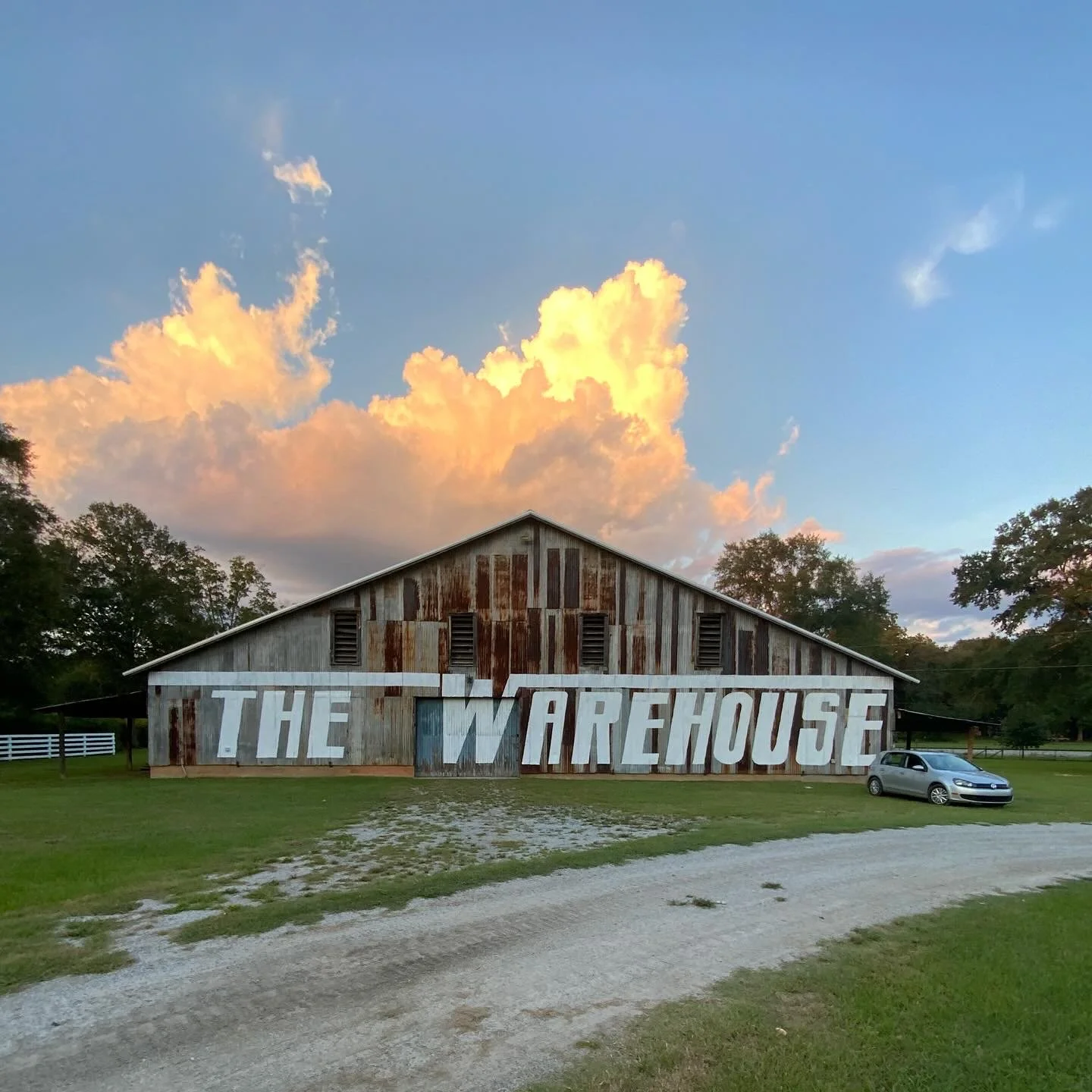 A rustic barn with a sign that reads 'The Warehouse' in large, spaced-out letters, parked on a grassy field with a gravel road and trees in the background, under a sky with dramatic clouds at sunset.