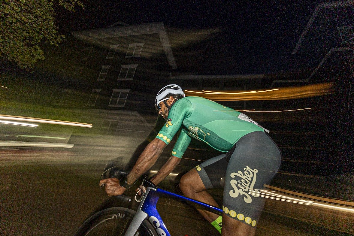 A cyclist in a green and black cycling outfit riding a bike at night with motion blur and light streaks in the background.