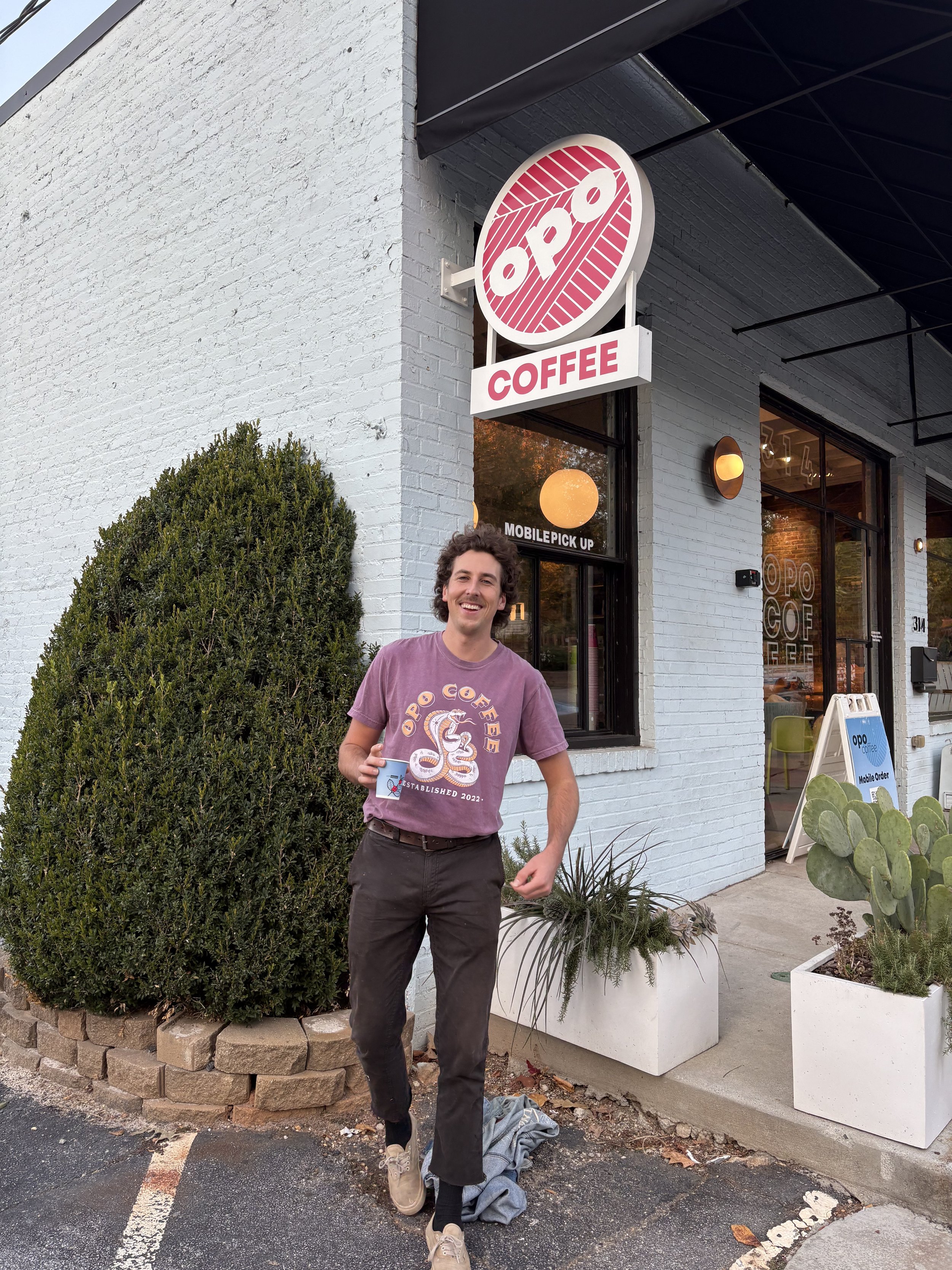 A man is walking out of a coffee shop called OPO Coffee, holding a coffee cup and smiling. The shop has a white brick exterior with a round sign displaying the name OPO and the word Coffee. There are plants in white planters outside, including a larg
