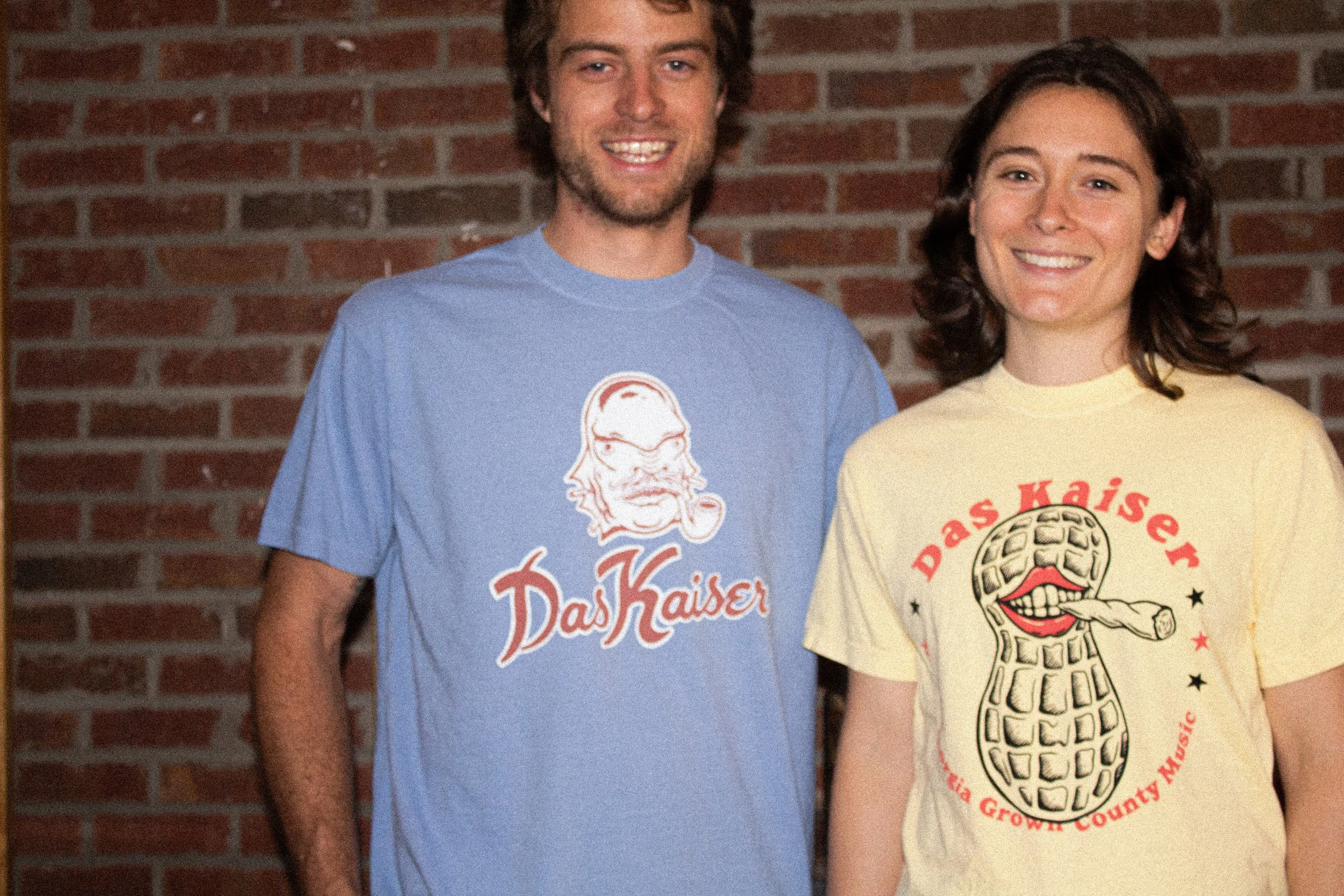 Two young adults, a man and a woman, are standing outside in front of a brick wall, smiling at the camera, wearing colorful t-shirts with graphics and text.