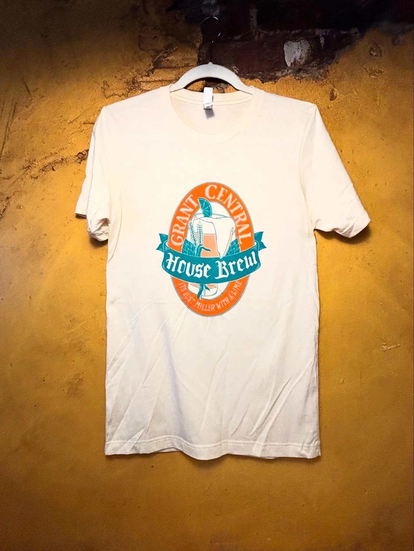 White t-shirt with a colorful logo design hanging on a hanger against a yellow wall. The logo features a beer glass with a lime, wheat, and hops, surrounded by orange and blue banners that read "Grant Central House Brew" and "It's just Miller with a 