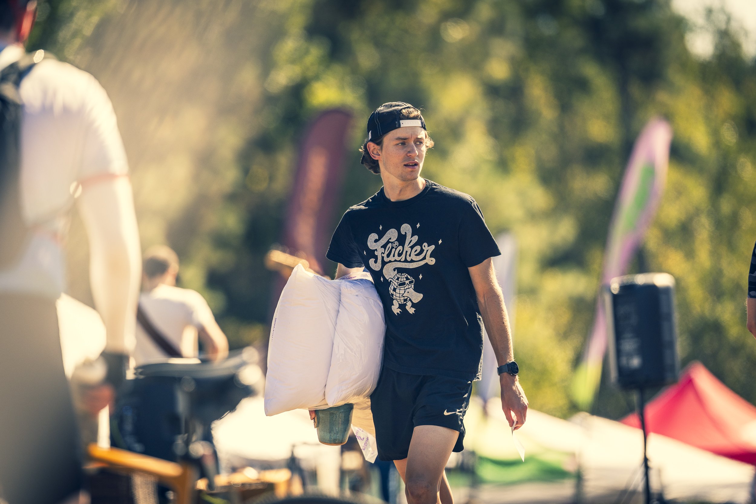 A young man wearing a black cap, black t-shirt, and black shorts walking outdoors carrying a pillow and a cup. There are other people and colorful banners in the background.