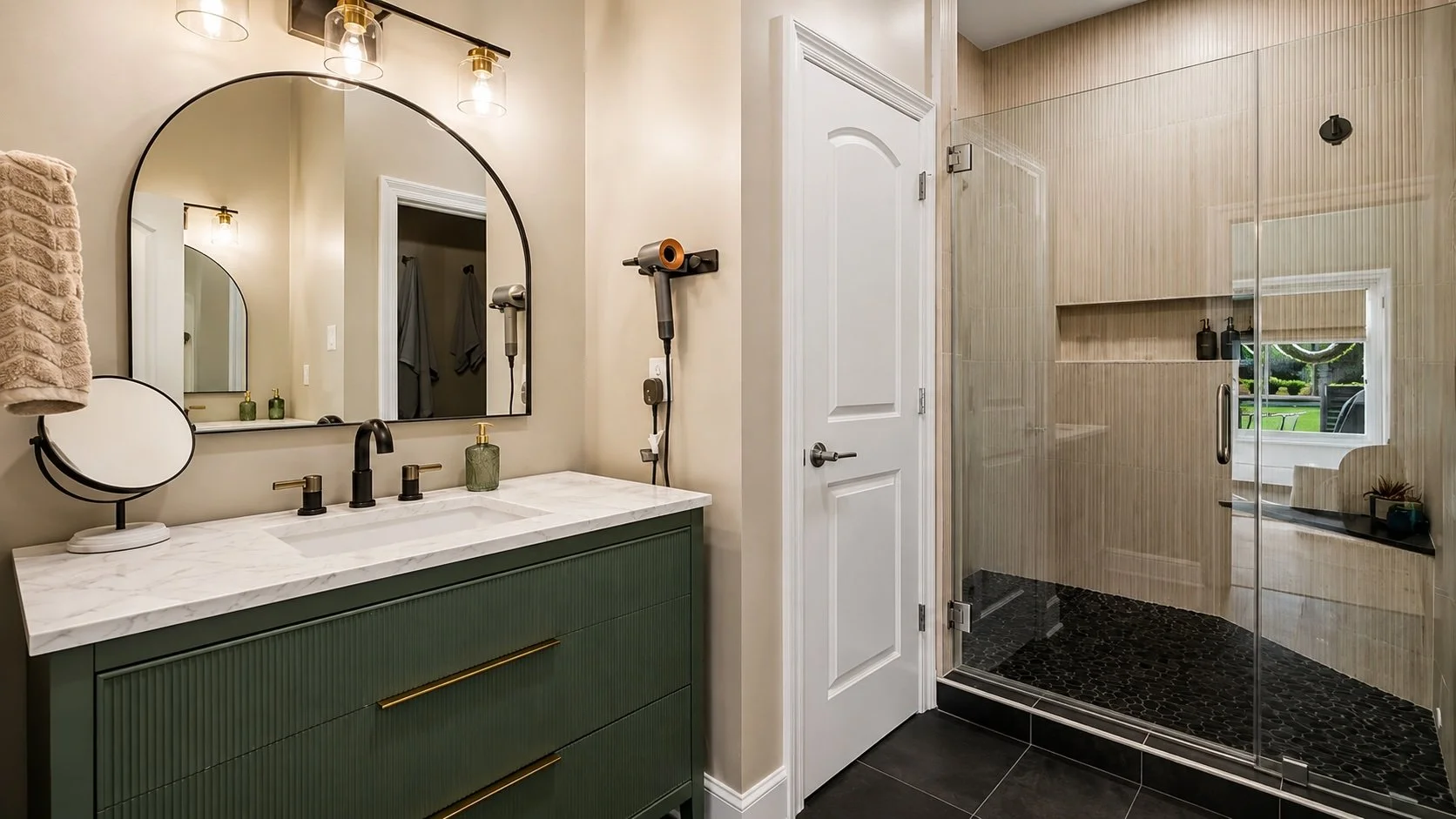 A stylish bathroom remodel completed in Covington, Louisiana, featuring a custom green vanity with marble countertops, brass accents, an arched mirror, and a spacious frameless glass shower. Textured wall tile and dark mosaic shower flooring add cont
