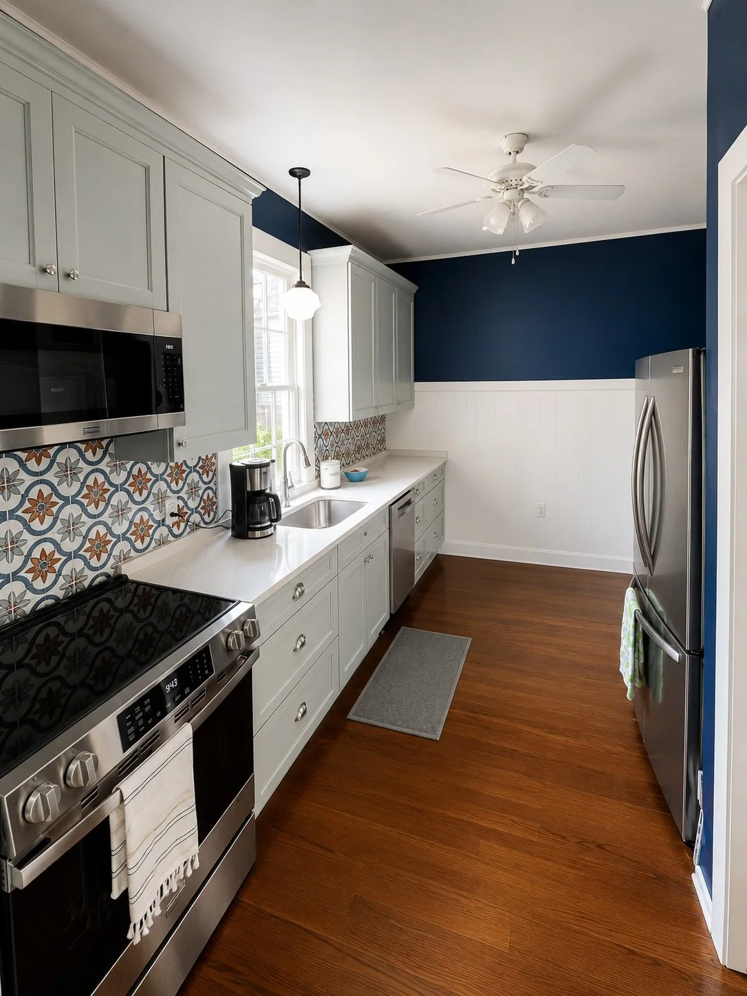This custom galley kitchen remodel by Grand Rising Construction showcases light gray shaker cabinets, quartz countertops, a decorative patterned tile backsplash, stainless steel appliances, updated lighting, and beautifully refinished hardwood floors