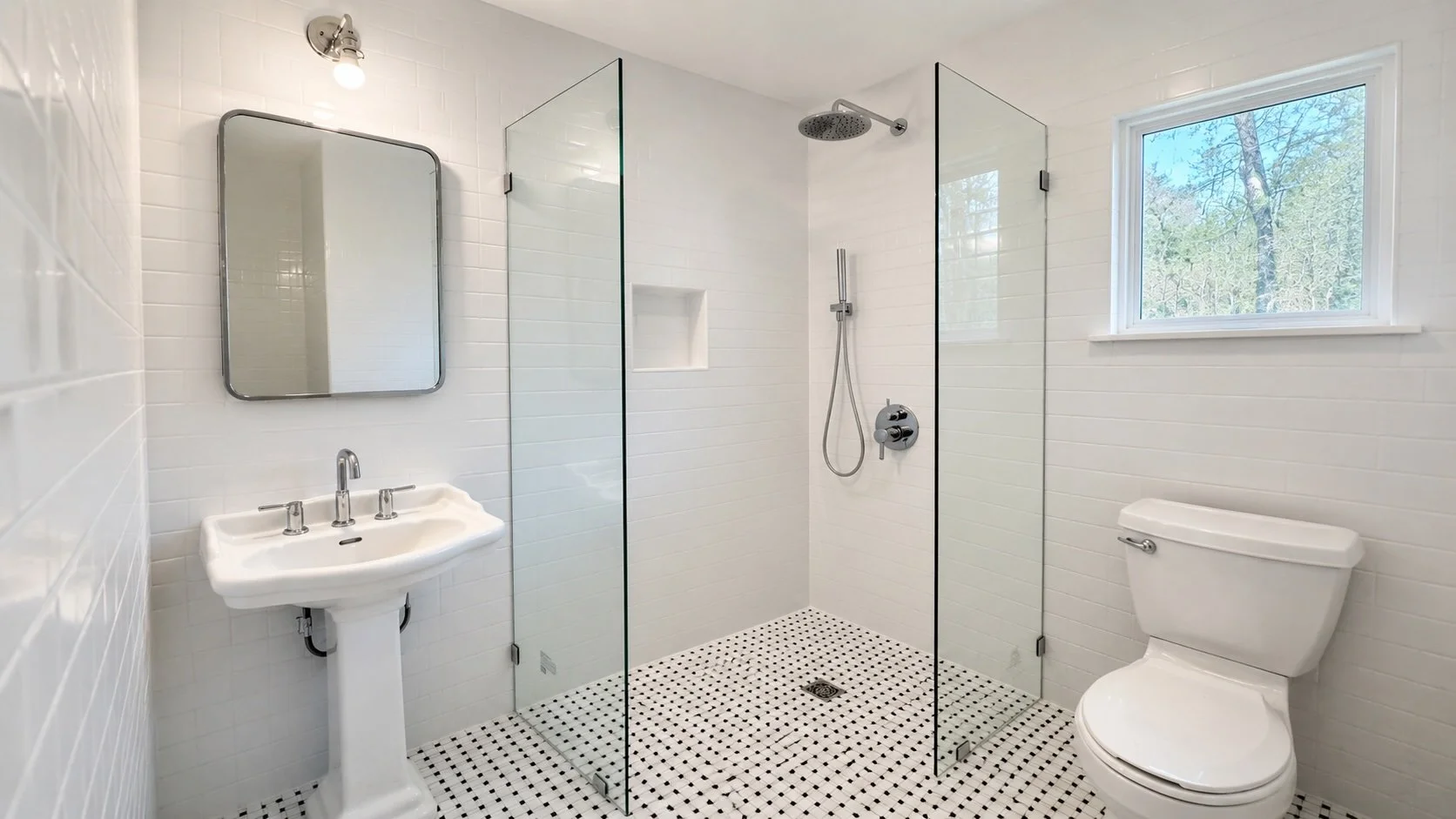 A clean, modern bathroom remodel completed in Goodbee, Louisiana, featuring a spacious walk-in shower with frameless glass, classic white subway tile, rainfall and handheld shower fixtures, a pedestal sink, and timeless mosaic flooring. The bright fi