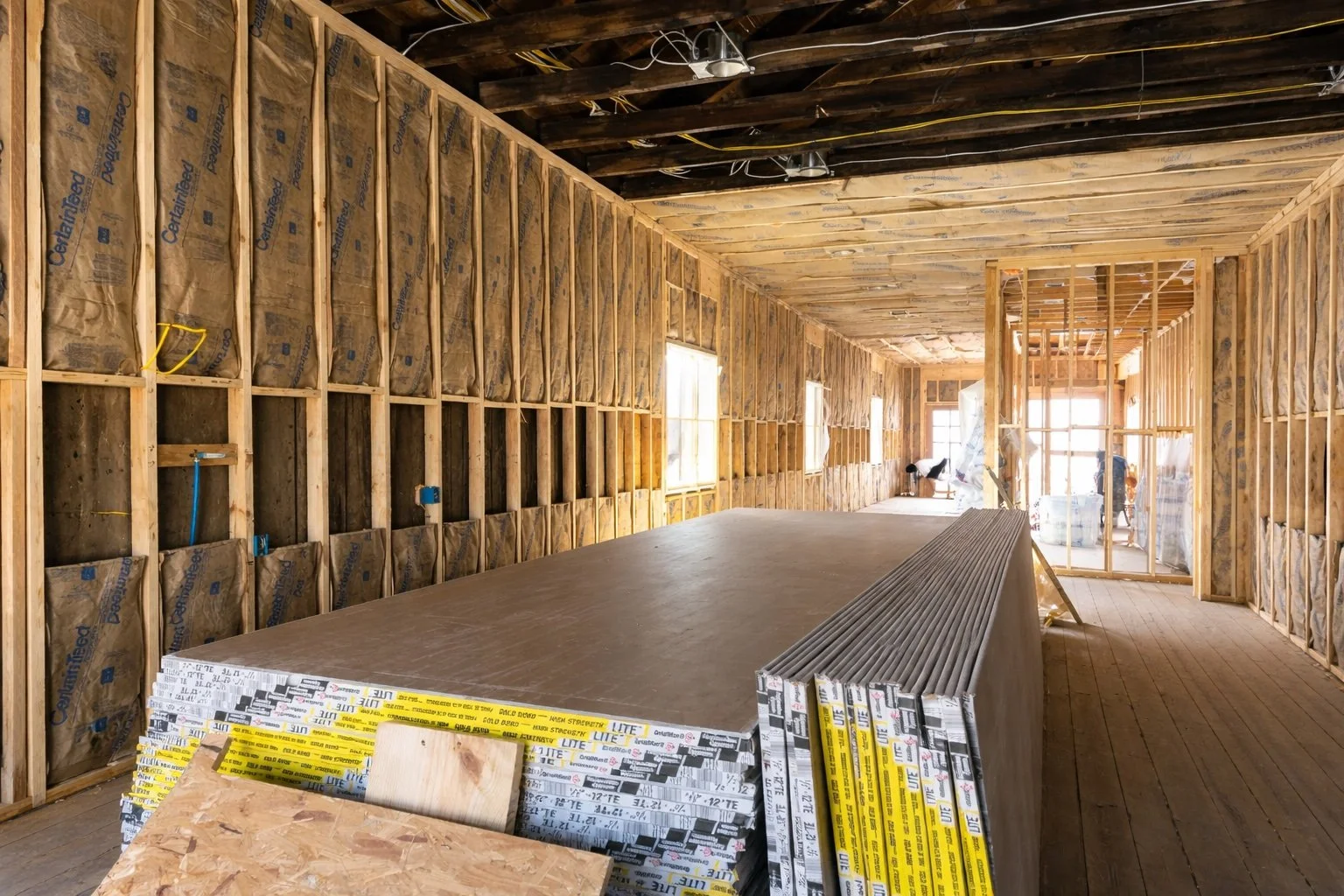 Wide-angle interior construction view showing framed walls with insulation and stacked drywall, brightened for a clean professional build-ready finish by Grand Rising Construction.
