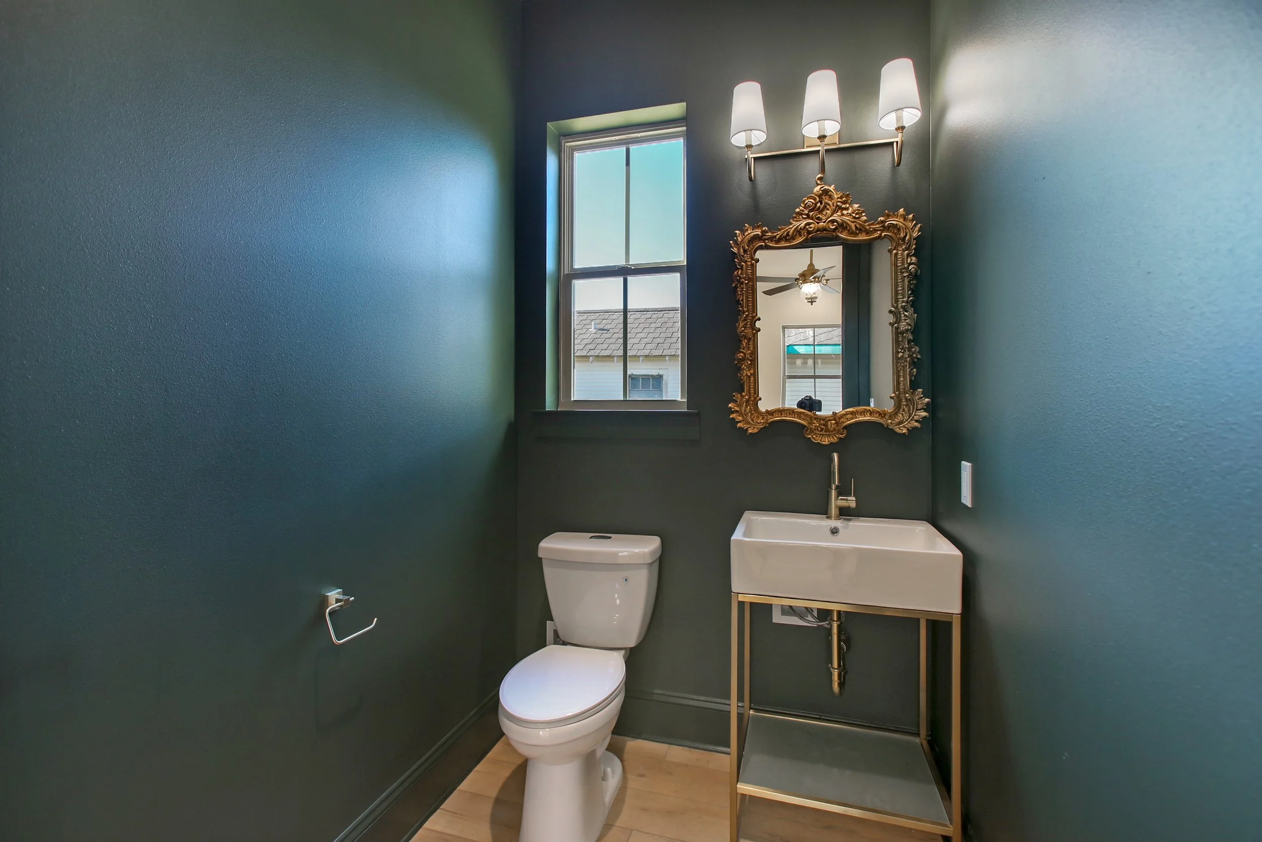 Elegant powder room remodel with dark green walls, gold ornate mirror, wall-mounted sink, and modern lighting.