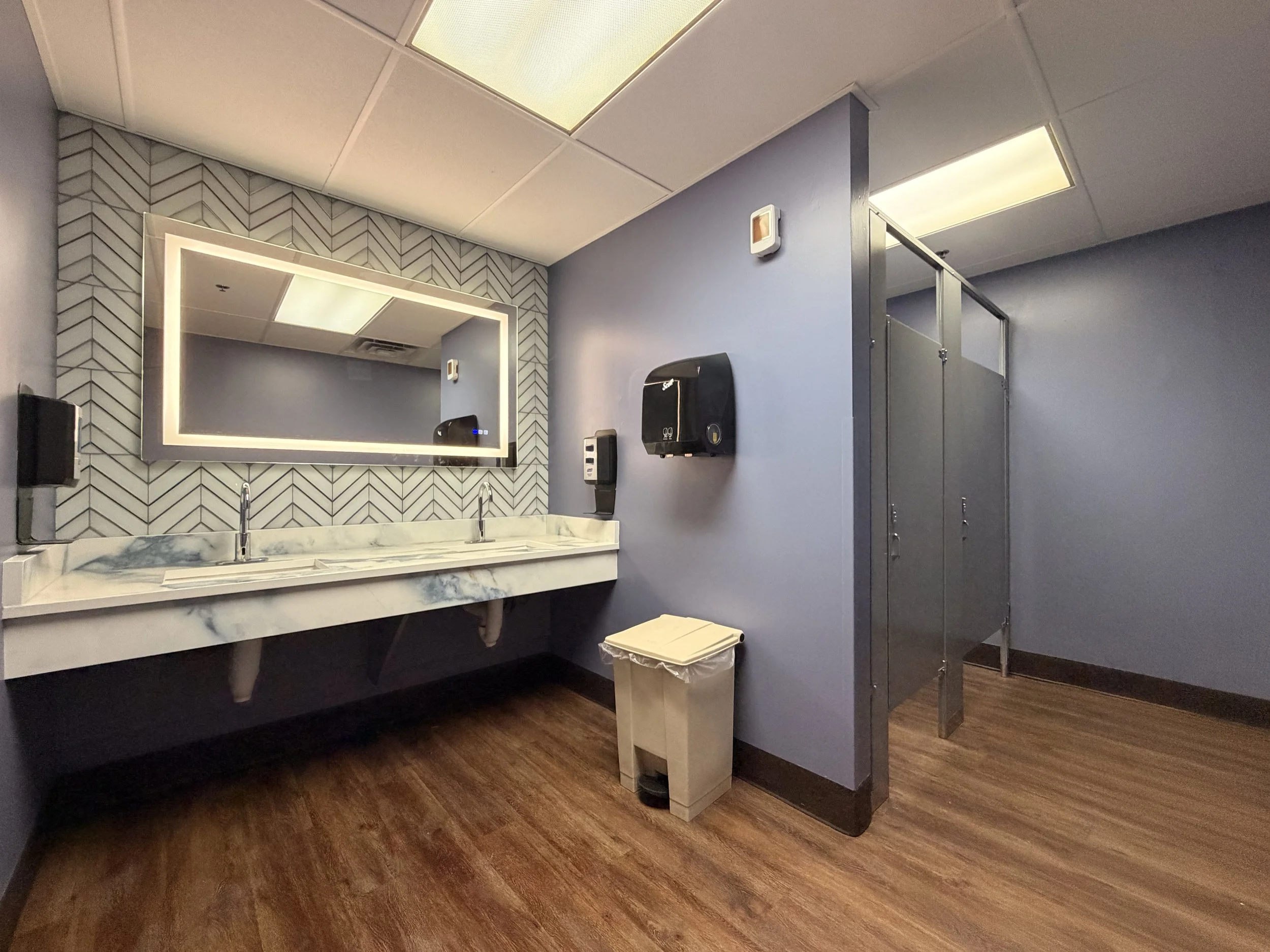 Grand Rising Construction completed this commercial bathroom remodel for a PAM Health facility in Covington, Louisiana. The renovation features a custom herringbone tile accent wall, illuminated LED vanity mirror, durable solid surface countertop wit