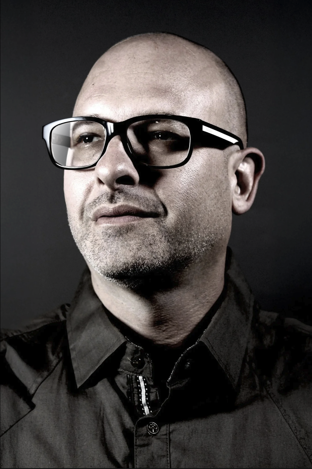 A man with a shaved head and glasses, wearing a black jacket against a dark background.
