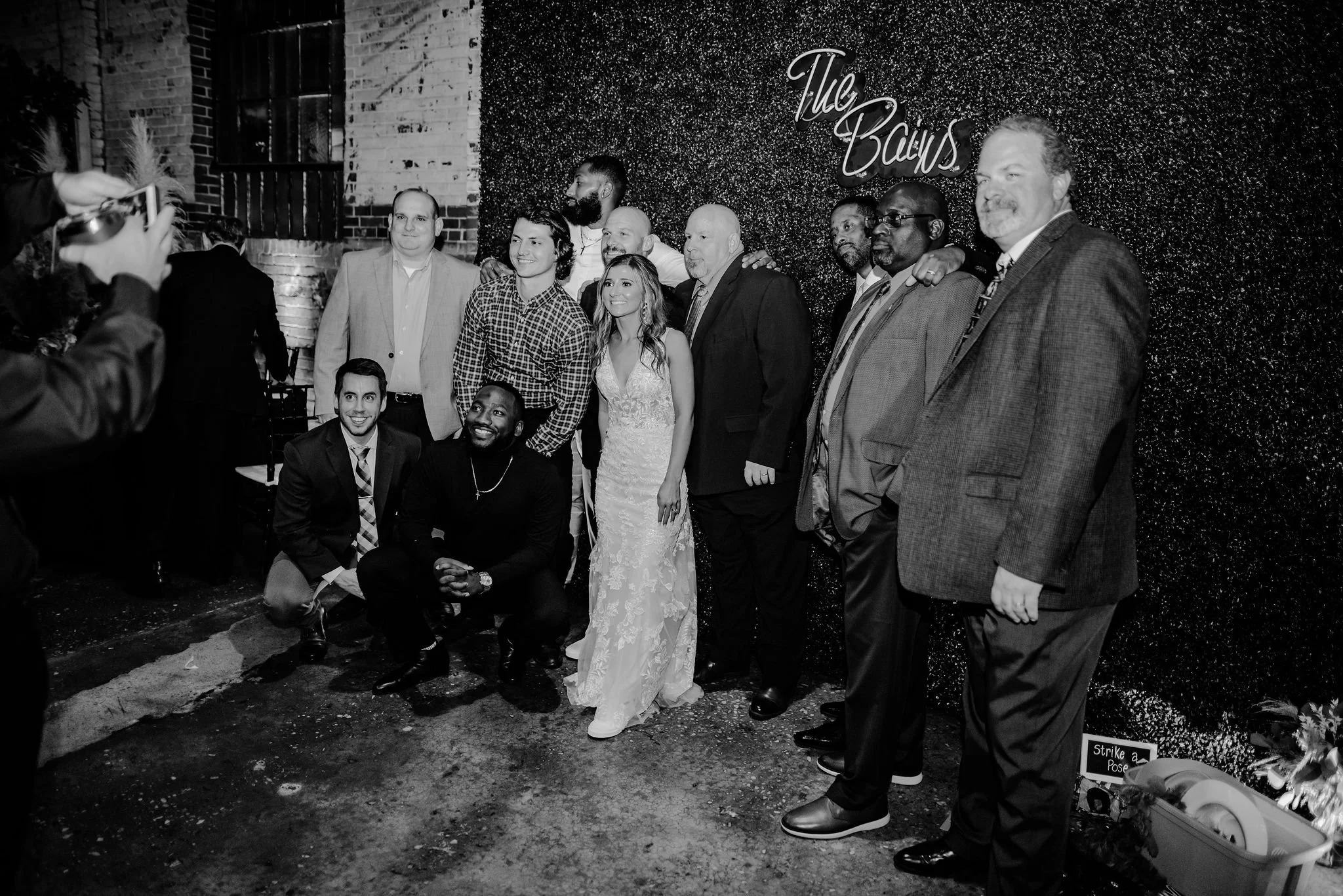 Group of people posing for a photo at a wedding reception, standing in front of a textured wall with the words "The Baus" written on it. Some are smiling, and a bride in a wedding dress is in the center.
