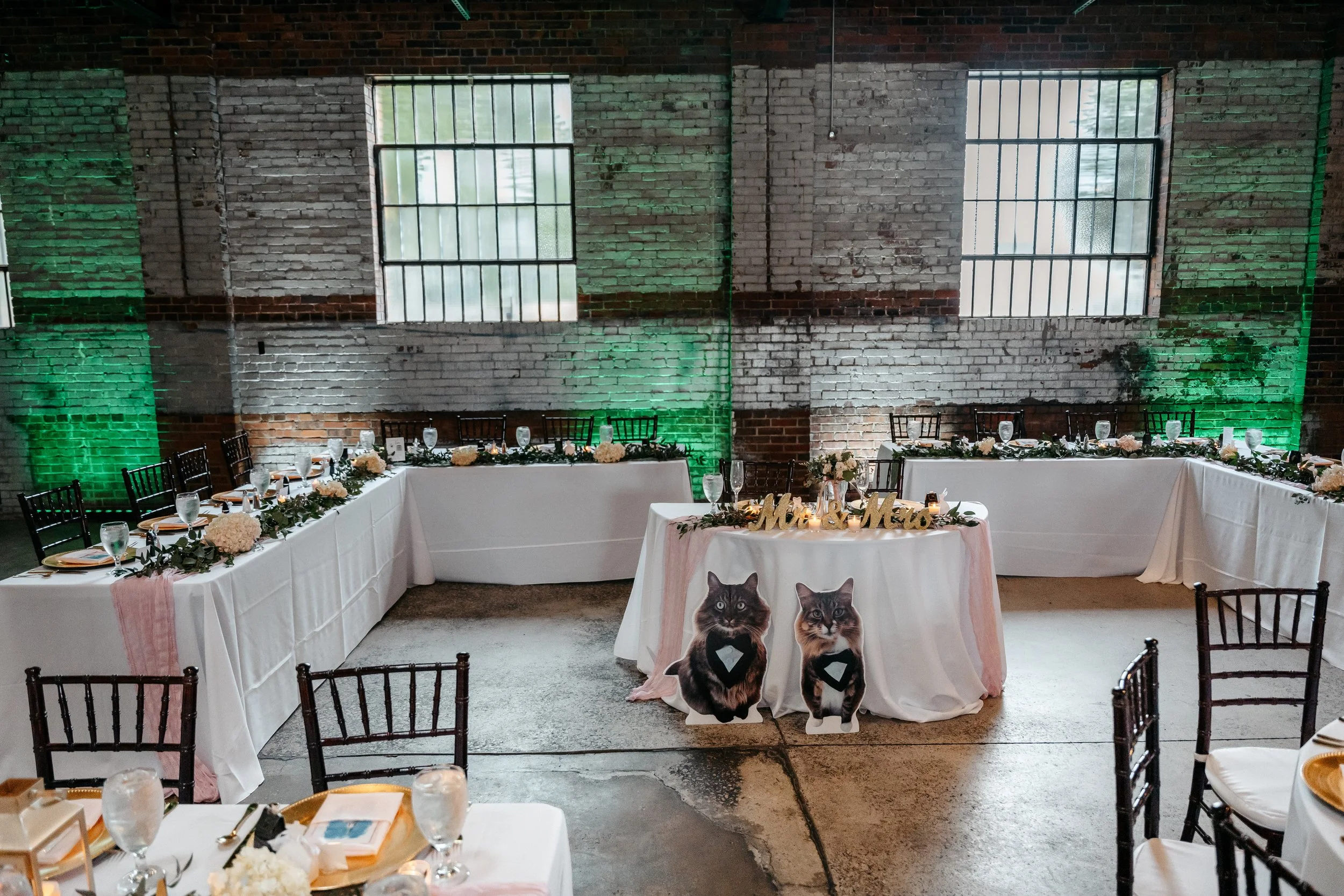 Interior of a wedding reception with U-shaped tables decorated with flowers and greenery, large photos of cats on what appears to be a cake table, and a sign that reads "Mr & Mrs" in gold letters. The room has exposed brick walls with windows and gre