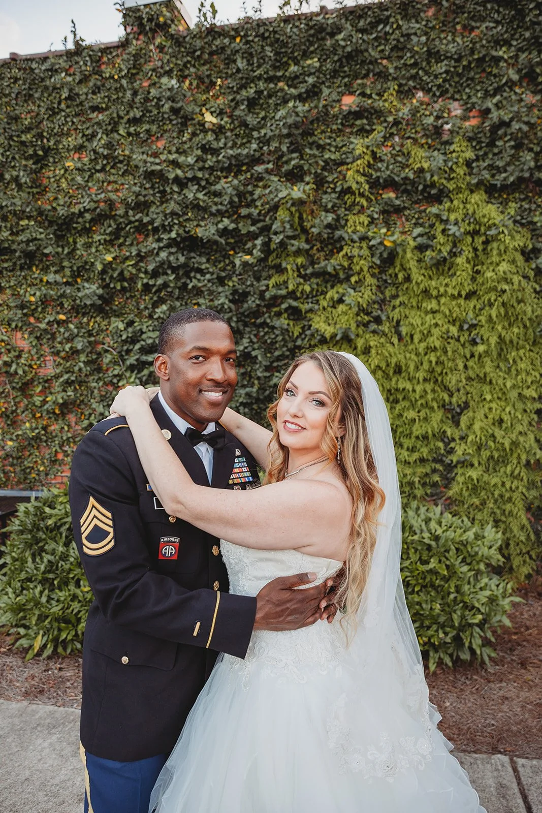 A couple, a bride in a white wedding gown and a groom in a military uniform, share a dance outdoors with a leafy green wall in the background.
