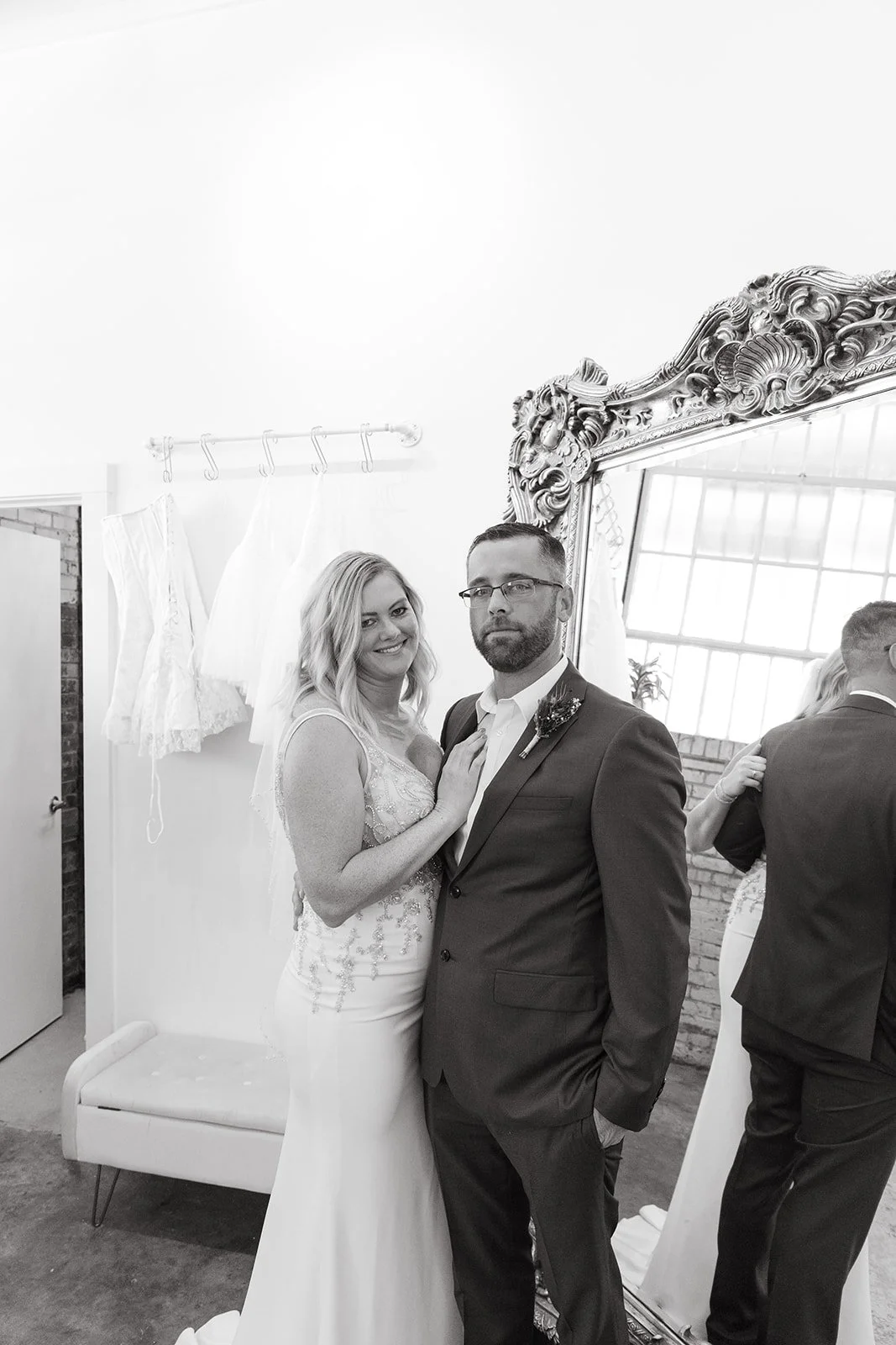 A bride and groom taking a mirror selfie, the bride wearing a lace wedding dress and the groom in a suit, in a room with a large ornate mirror and a window with grid panes.