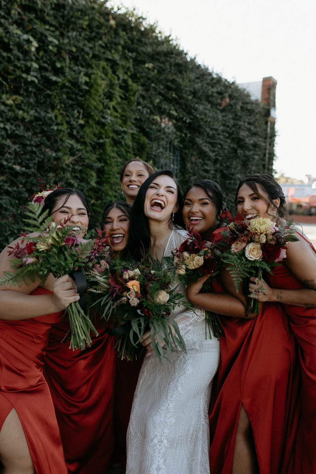 A bride and five women in red dresses holding bouquets, smiling and laughing outdoors in front of a green hedge.