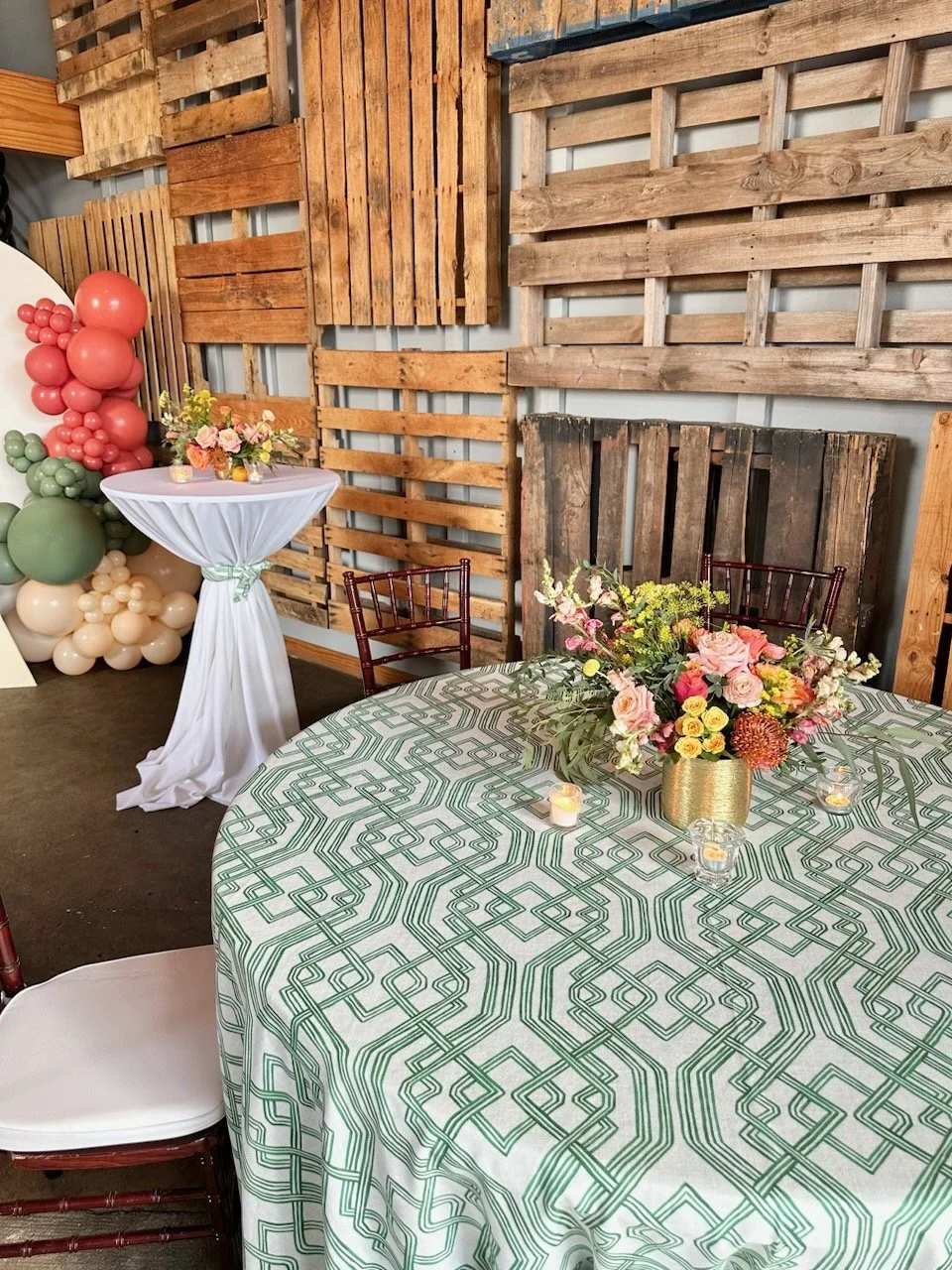 Decorative event setup with a round table covered with a geometric patterned tablecloth, a large floral centerpiece in a gold vase, small candles, and a wooden pallet backdrop, alongside a high-top table with a floral arrangement and balloon decoration.
