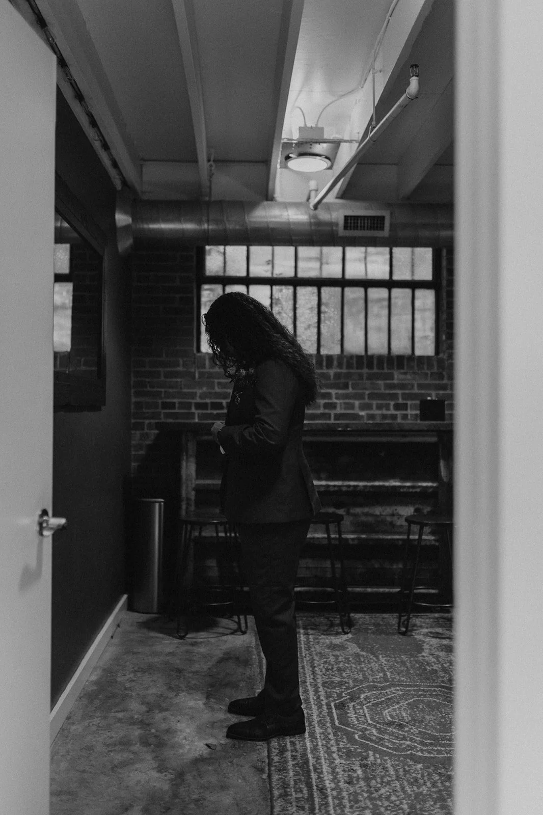 A woman with curly hair wearing a blazer standing in an industrial-style room with brick walls and large windows, looking down at her phone.