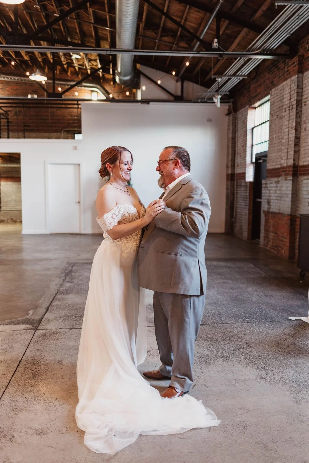 A bride and an older man, possibly her father, dance and smile at a wedding reception in an industrial-style venue with exposed brick walls and large windows.
