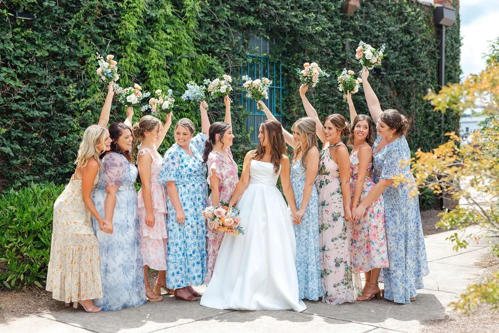 Group of women, including a bride in a white wedding dress, standing outdoors in front of greenery, celebrating with bouquets and smiling.
