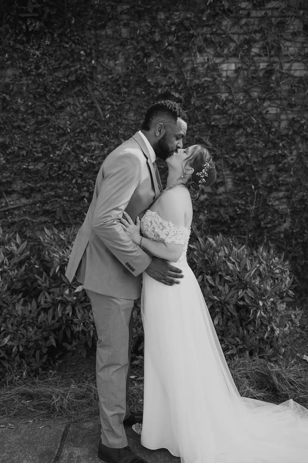 A black-and-white photo of a couple in wedding attire sharing a kiss outdoors, with a leafy background.