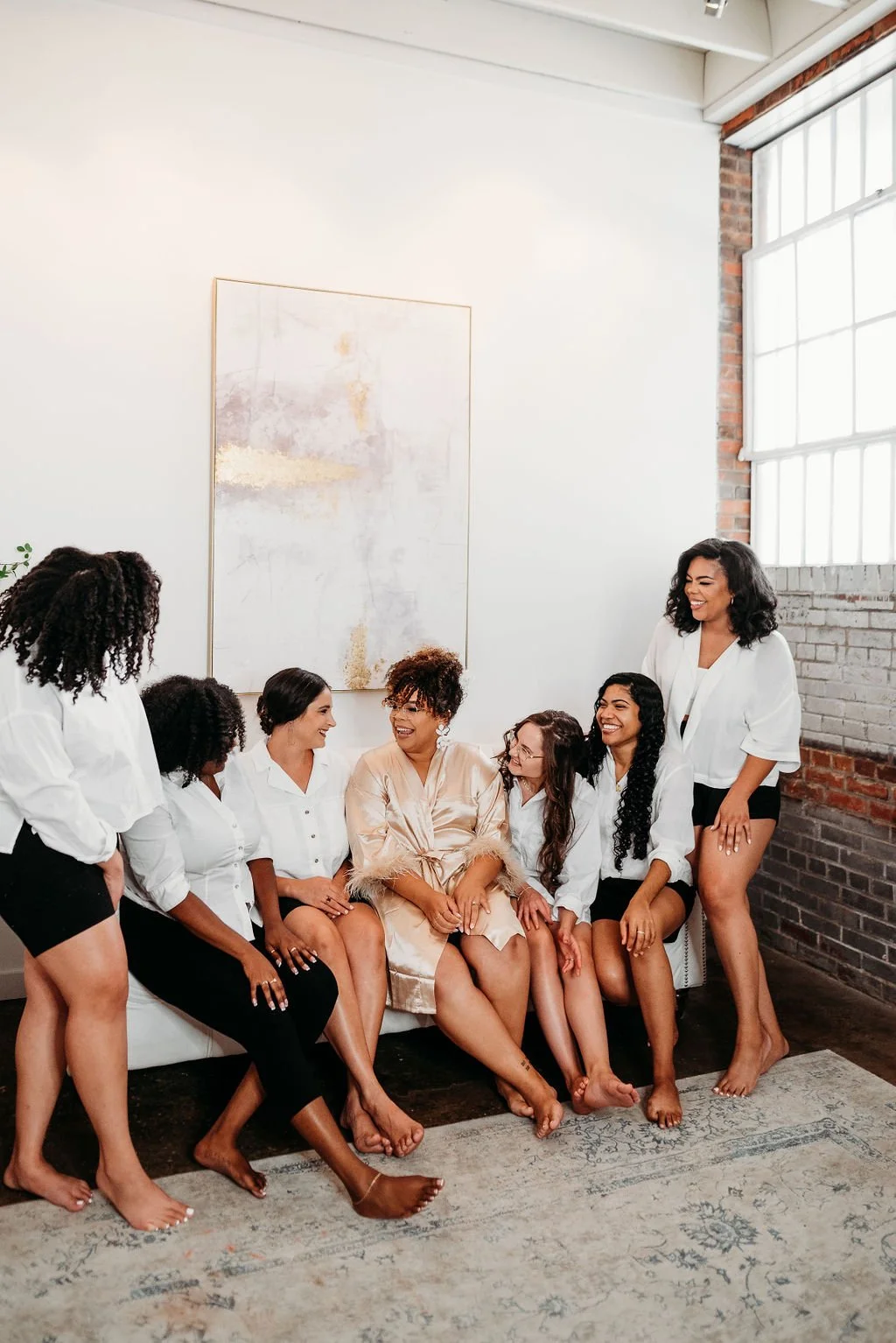 A group of women sitting and standing together in a bright room, smiling and laughing. They are dressed in white shirts and black shorts or pants, with one woman in the middle wearing a satin robe. The room has large windows, a white wall with artwork, and a brick wall.