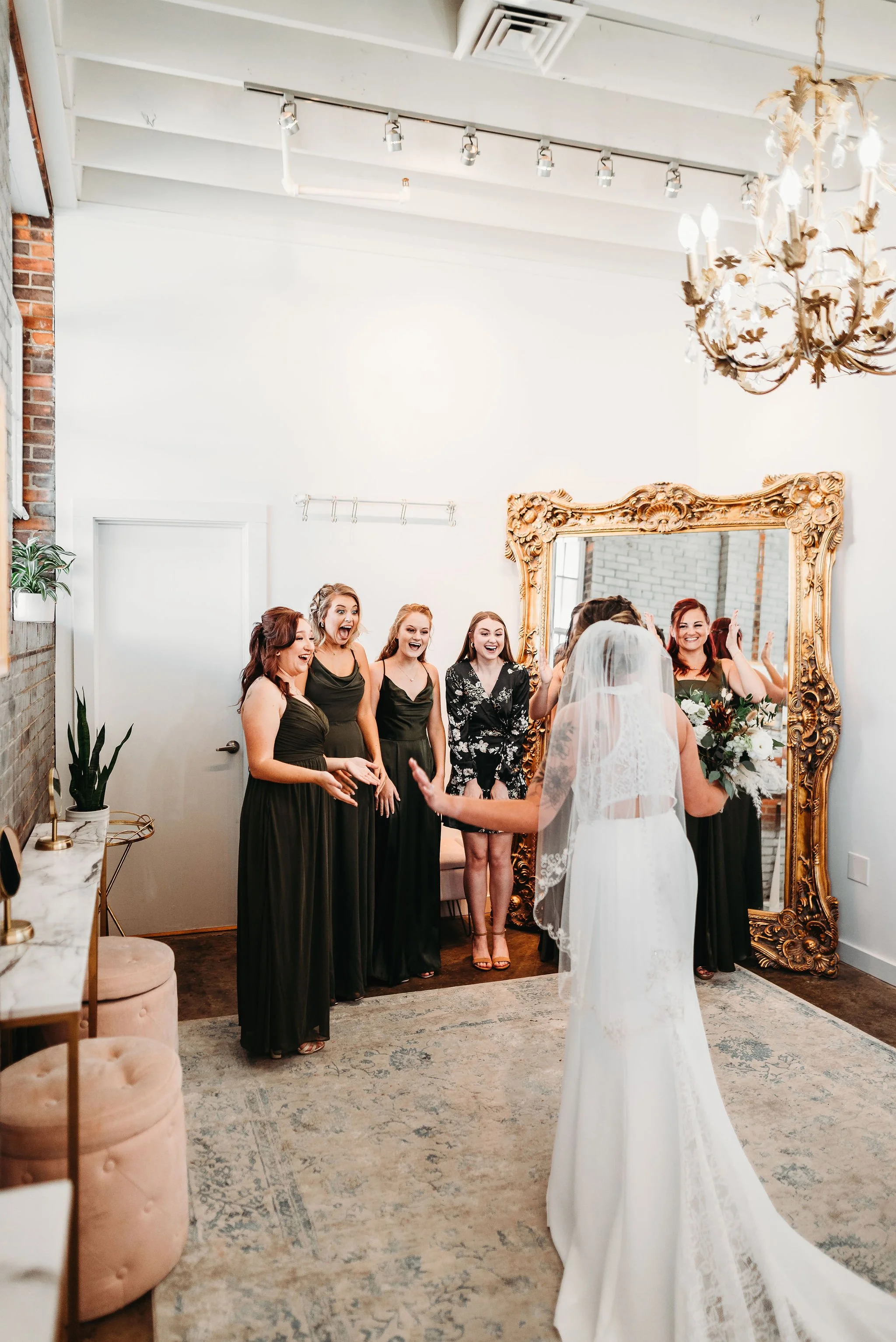 A bride with a wedding gown and veil faces five women in black dresses, smiling and reacting happily in front of a large ornate mirror in a bright, decorated room.
