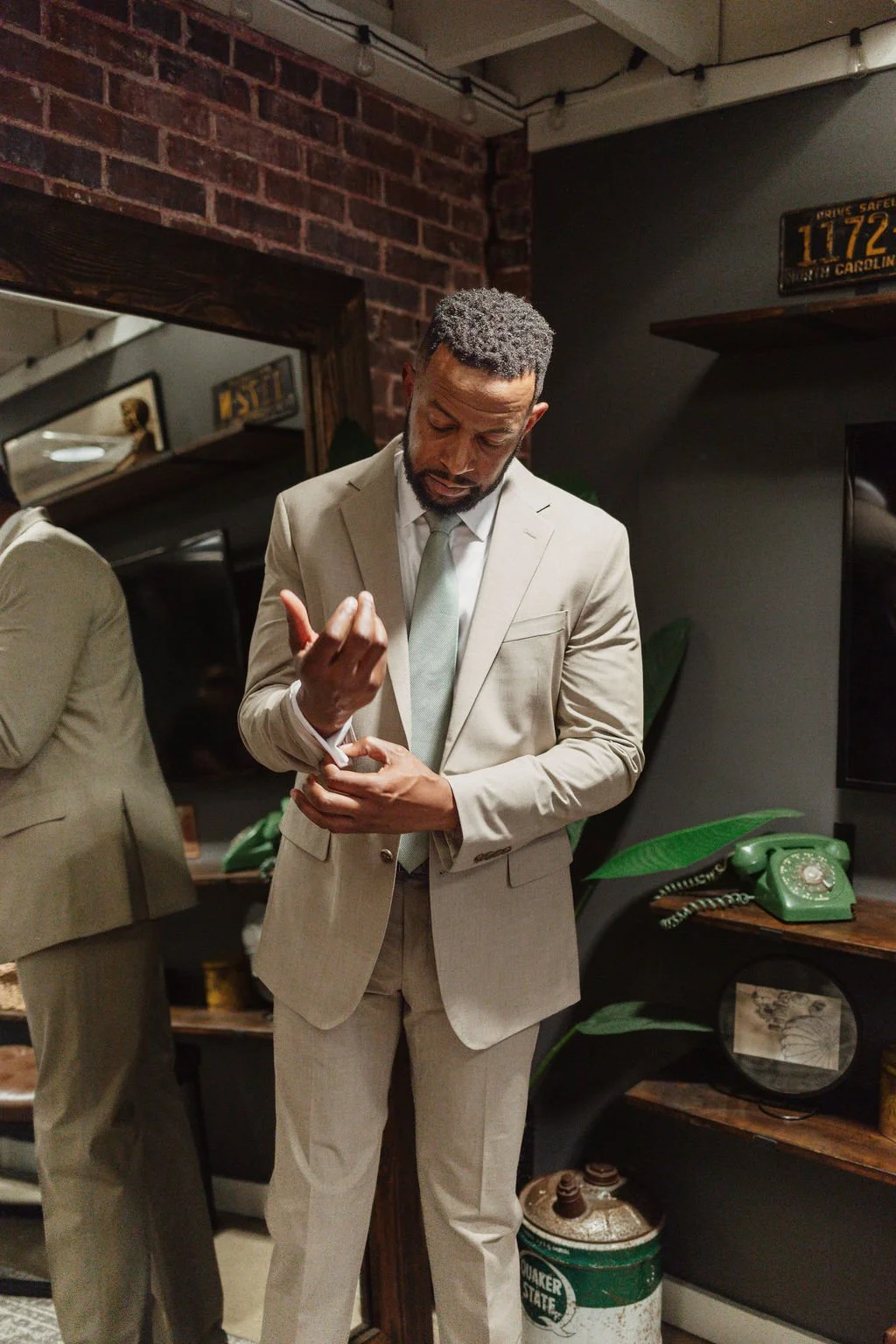 A man in a beige suit looking at his watch in a vintage-style room with brick wall, shelves, and a green rotary phone.