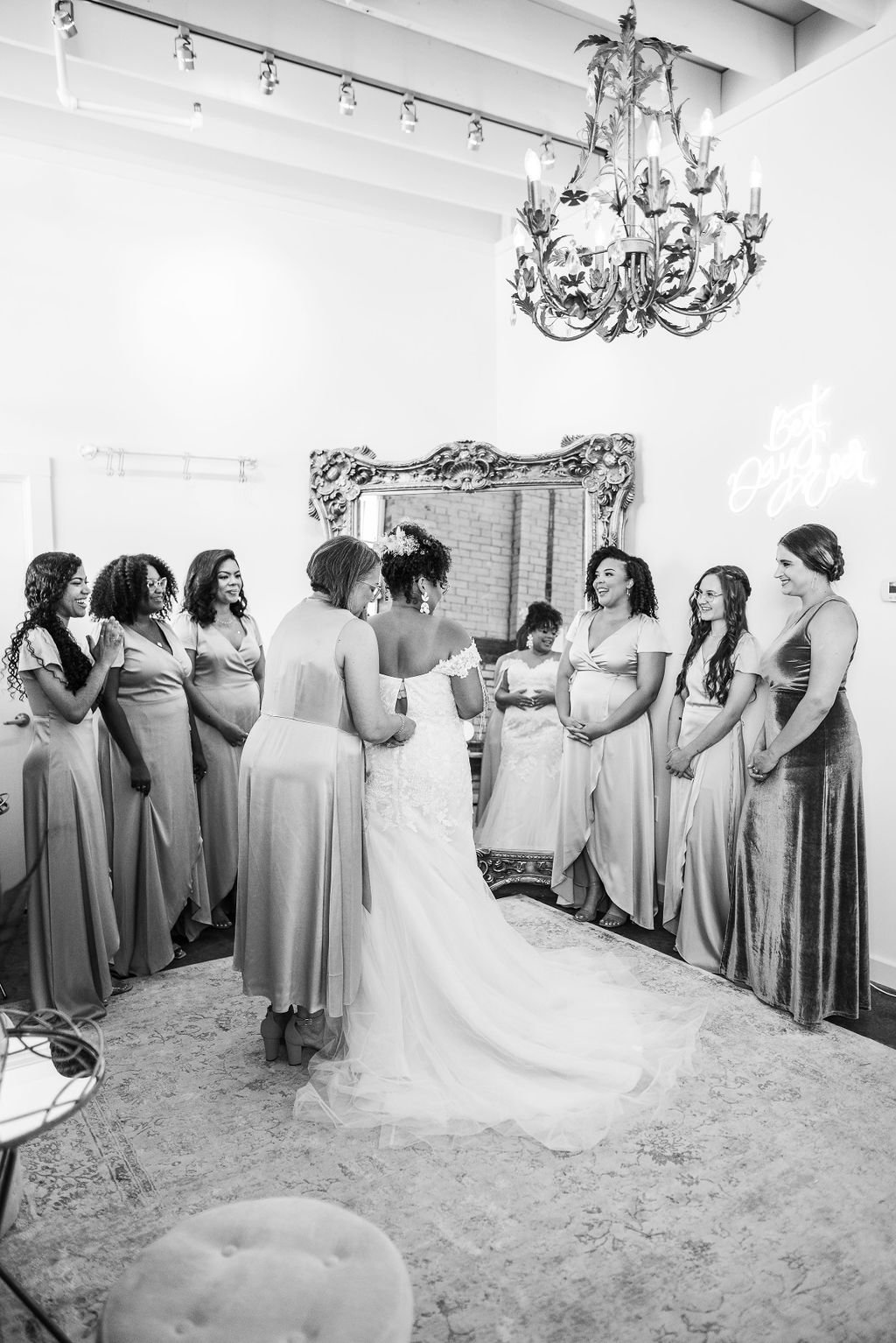 A bride in a wedding dress stands with her bridesmaids and friends in a room decorated with a chandelier and a large ornate mirror, as they prepare for a wedding.