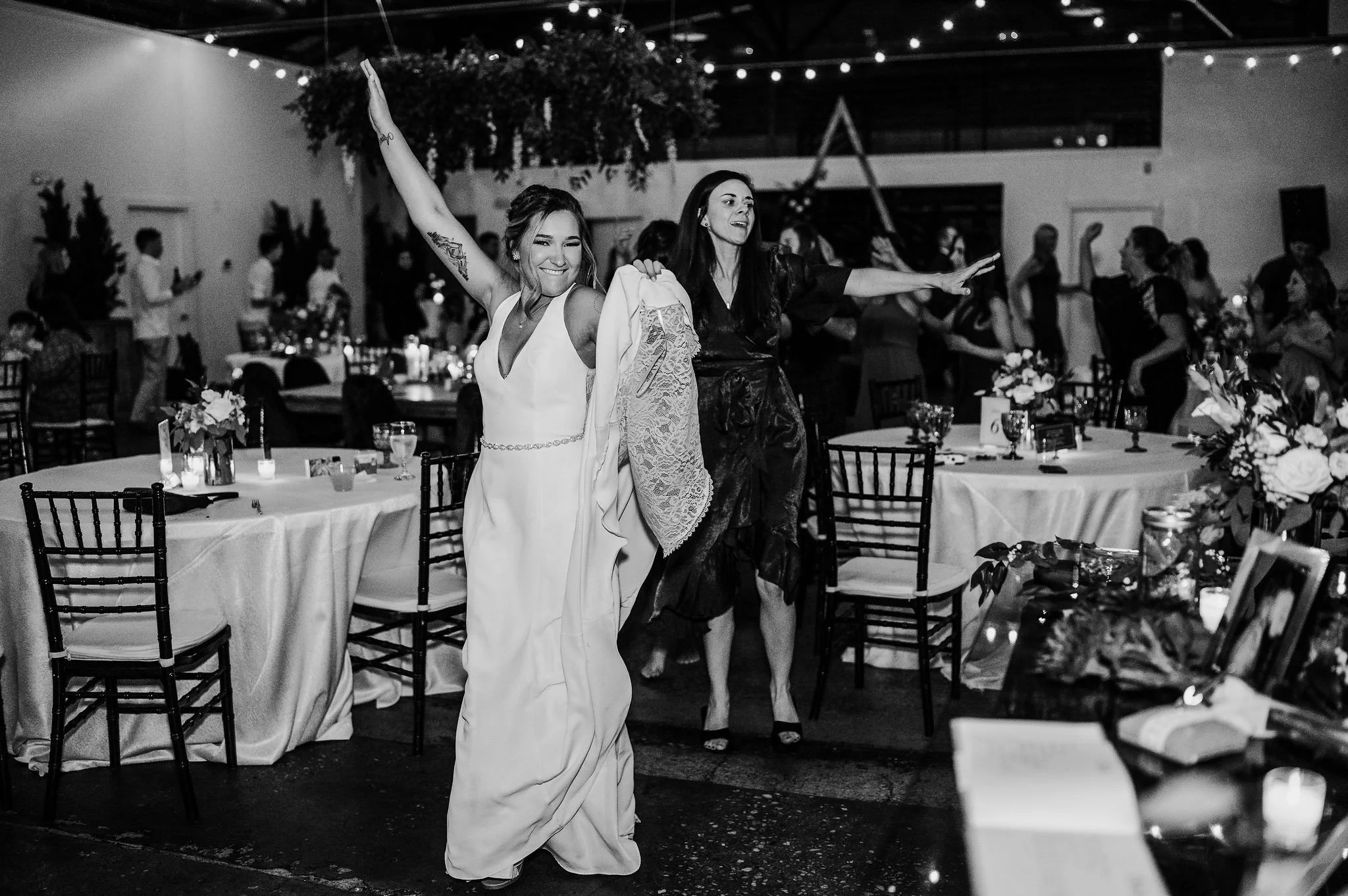 Women dancing at a wedding reception with tables, floral centerpieces, and guests in the background.