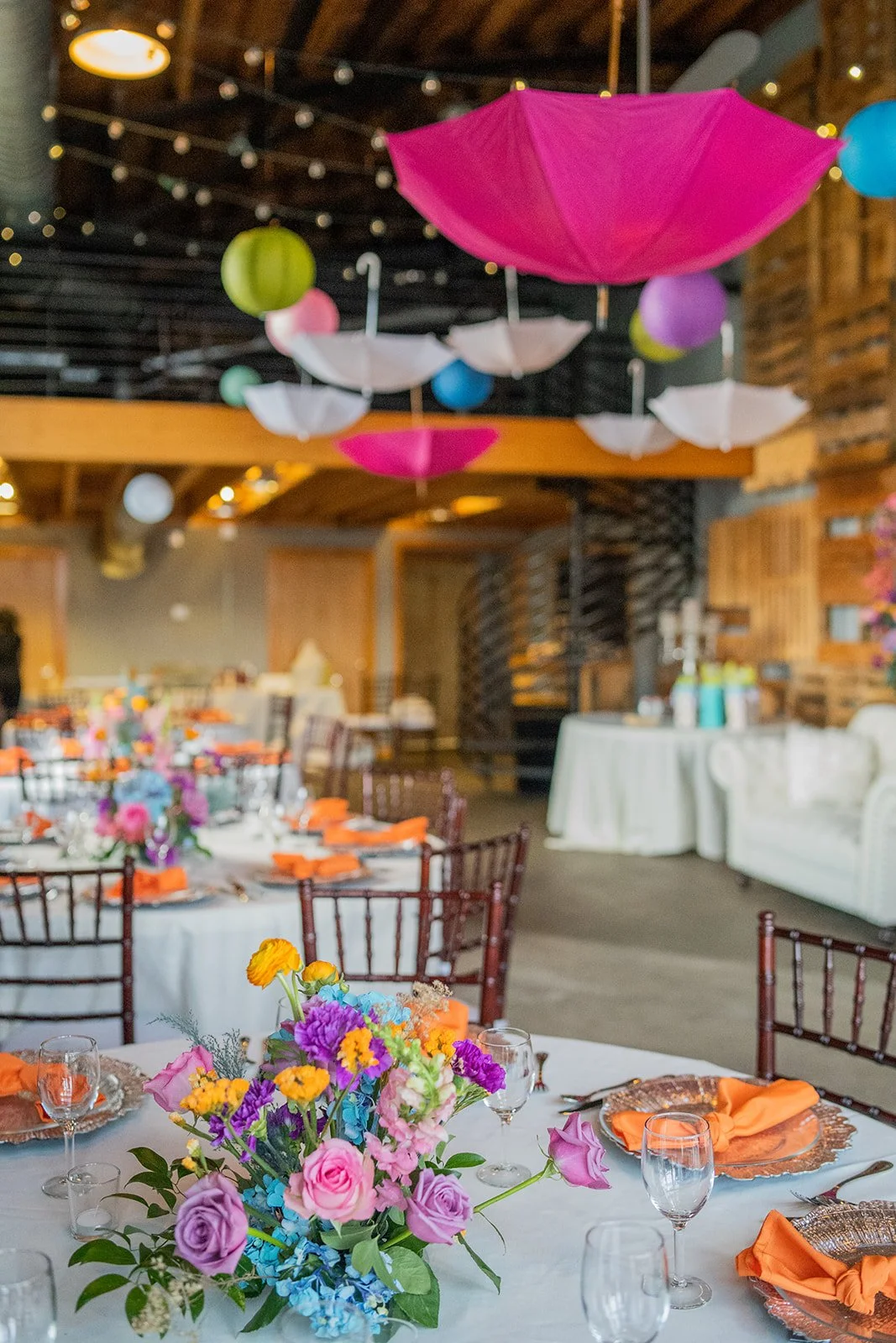 Decorated indoor event space with hanging paper umbrellas and lanterns, round tables with floral centerpieces, glassware, and table settings, cozy seating area with a white couch, and warm wooden interior.