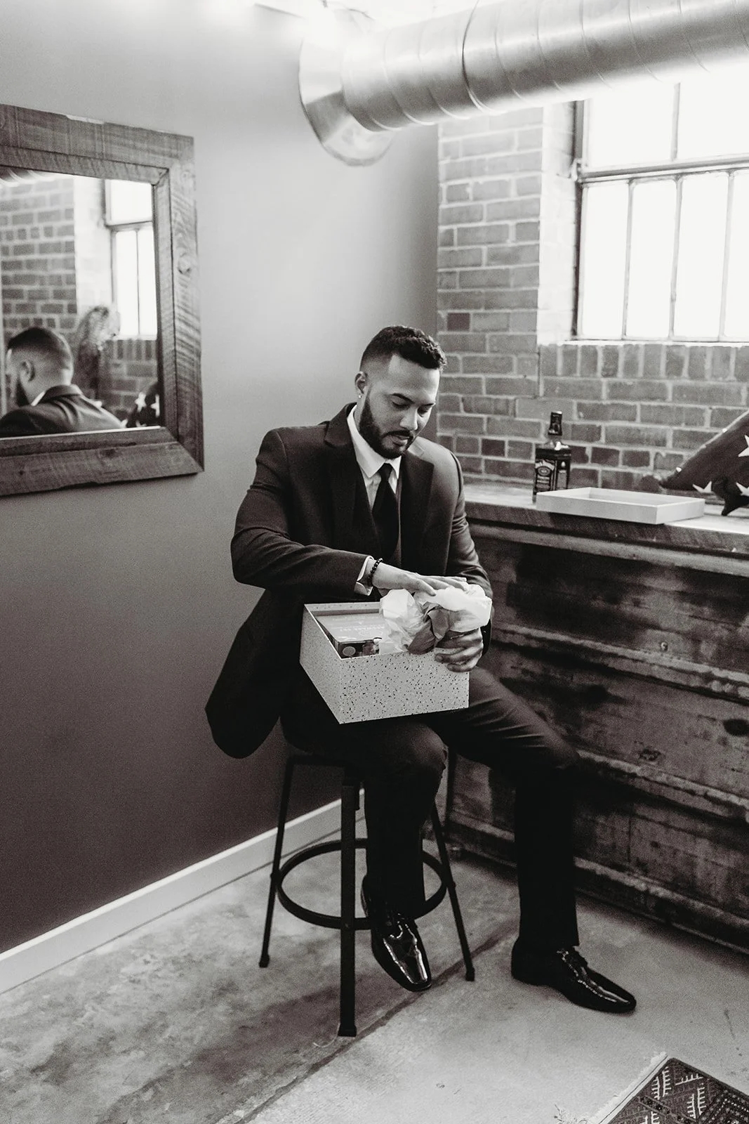 A man in a suit sitting on a stool opening a gift box in a room with brick walls and a window, with a mirror reflecting his back.