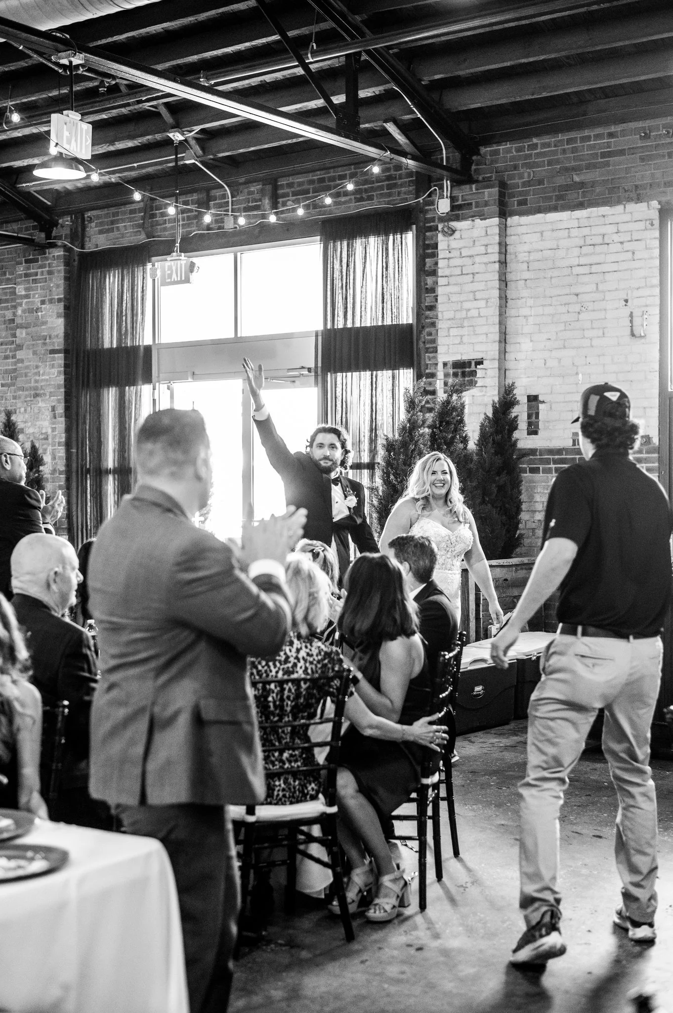 Black and white photo of a wedding reception with a couple on stage. The groom is raising his hand while the bride is smiling. Guests are seated and clapping, with some taking photos. Decorations include string lights and potted plants inside a rusti