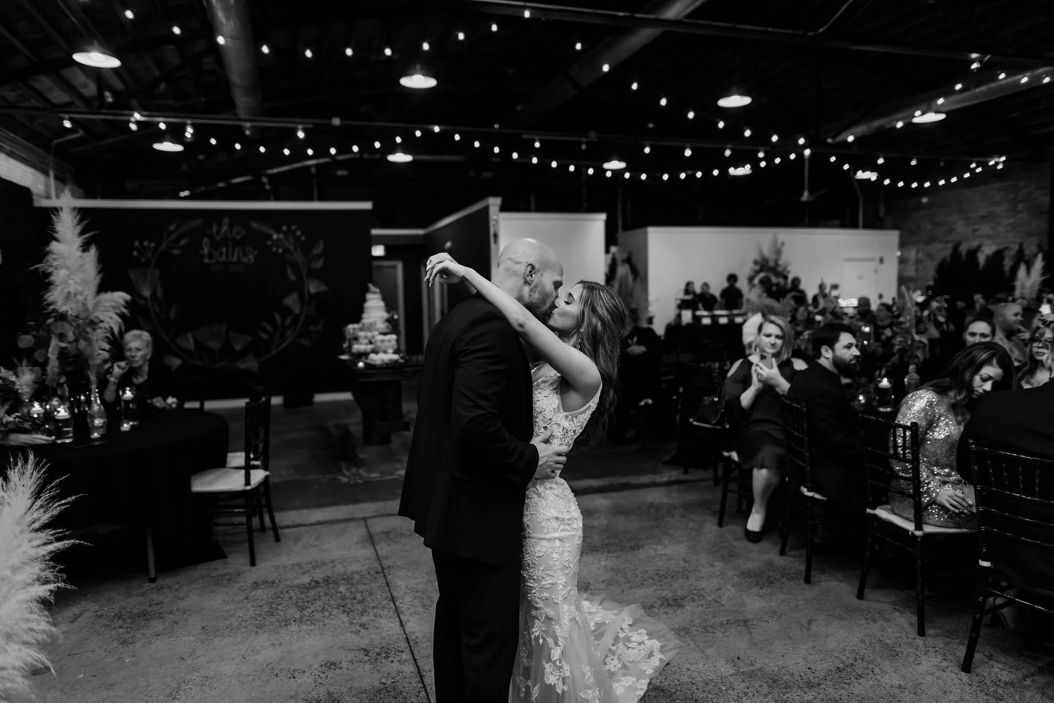 A couple sharing a dance at their wedding reception, with guests seated at tables behind them and string lights overhead.