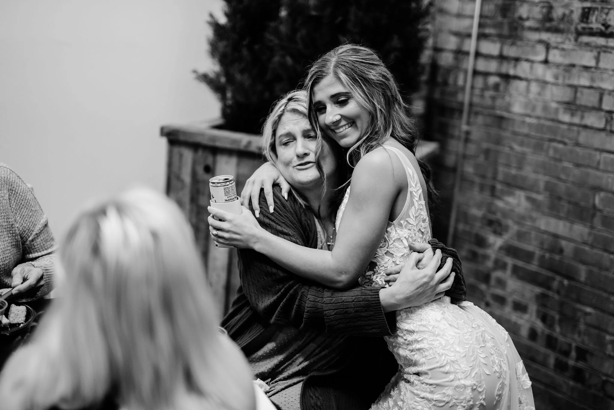 A woman in a wedding dress hugging and smiling with an older woman at a social gathering. The scene is in black and white.