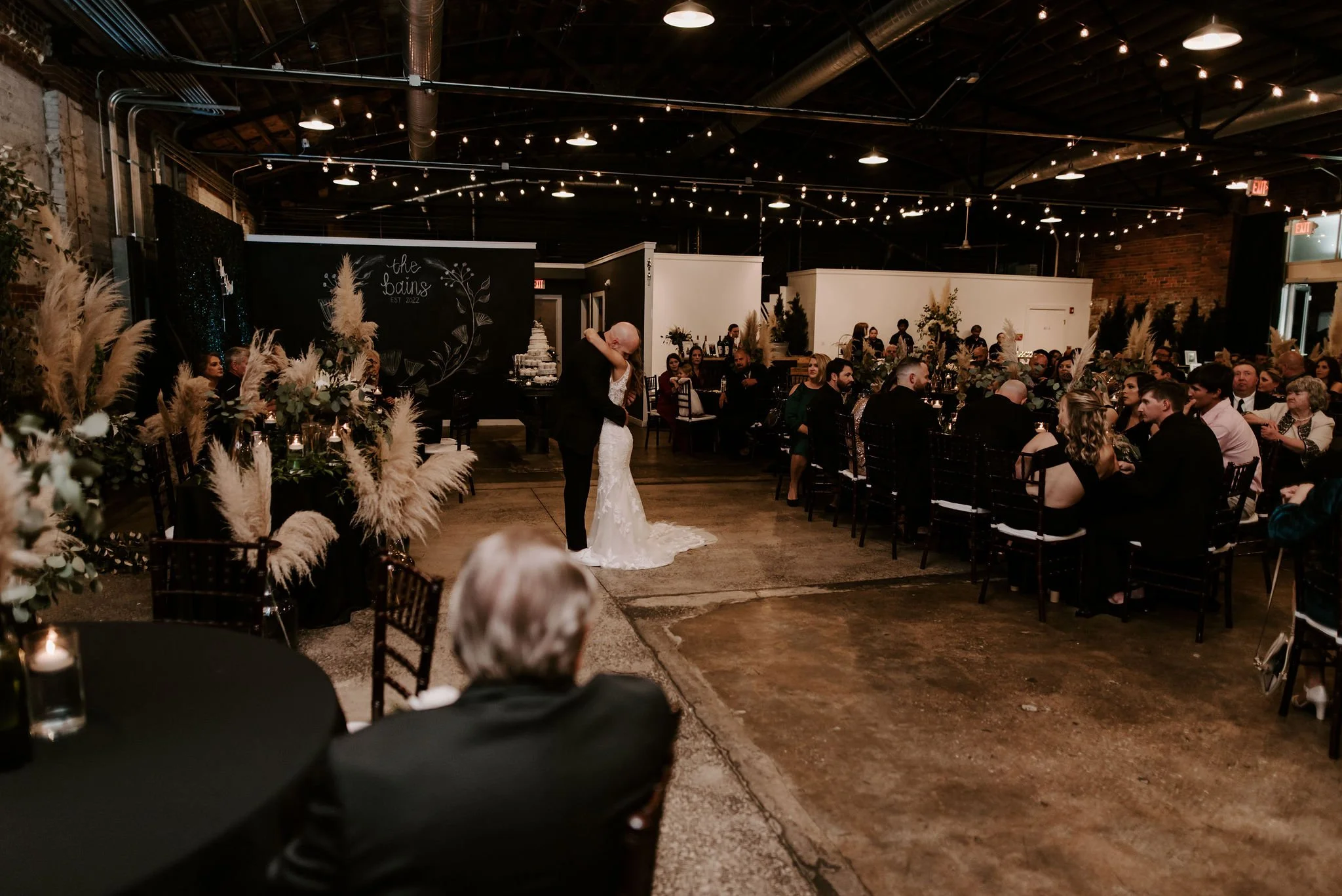 Indoor wedding reception with a bride and groom sharing a dance, surrounded by seated guests, decorated with floral arrangements and string lights.