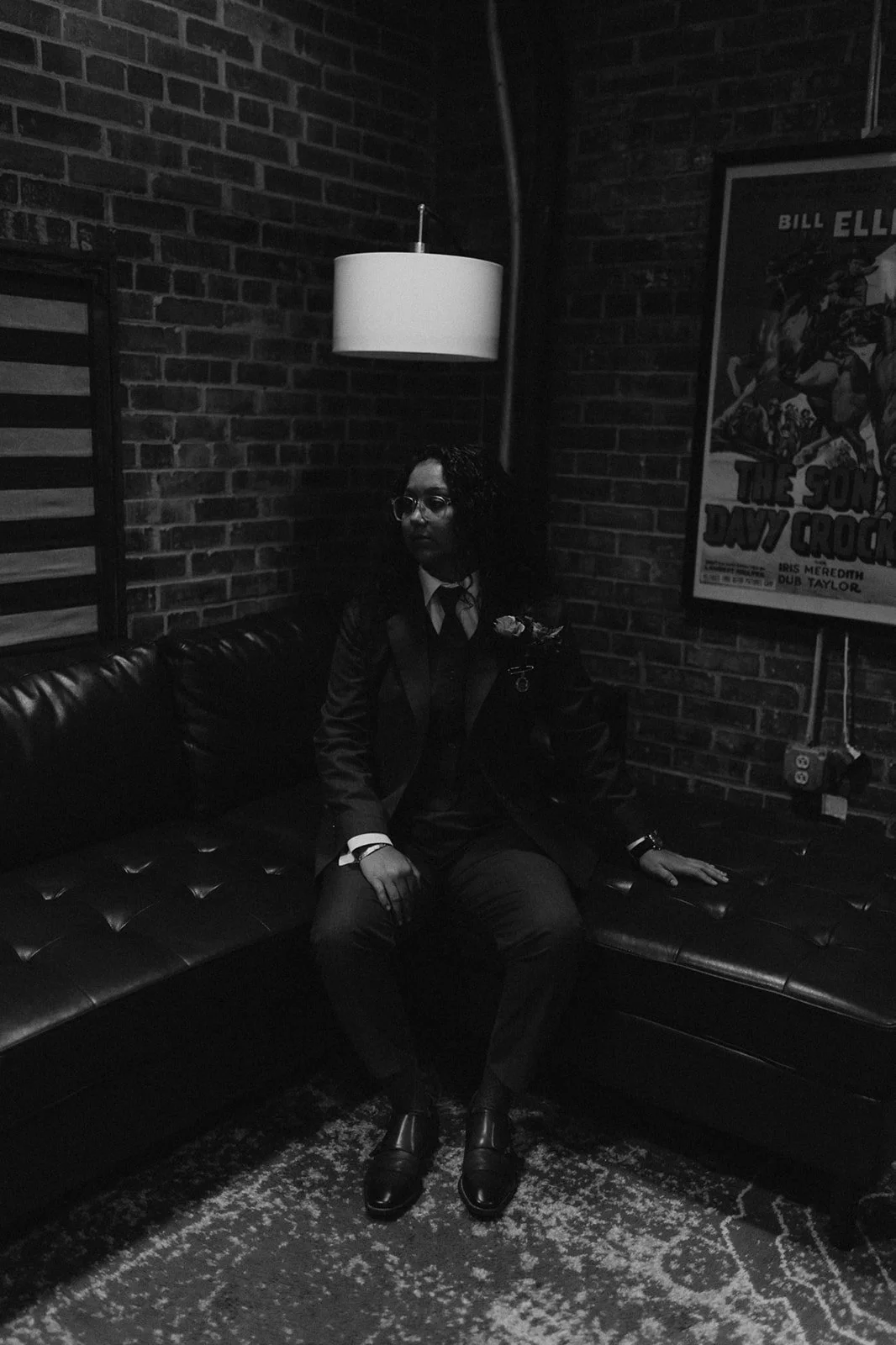 A person dressed in a formal, dark suit with a tie, sitting on a leather sofa in a dimly-lit room with brick walls, a hanging lamp, and a vintage movie poster.