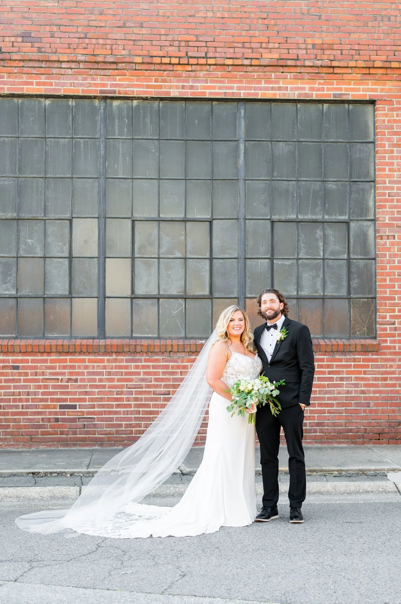 A newlywed couple stands on the street in front of a large brick wall with a grid of old, weathered industrial windows. The bride wears a white wedding gown with lace detail, holding a bouquet of white flowers and greenery, with a long veil flowing b