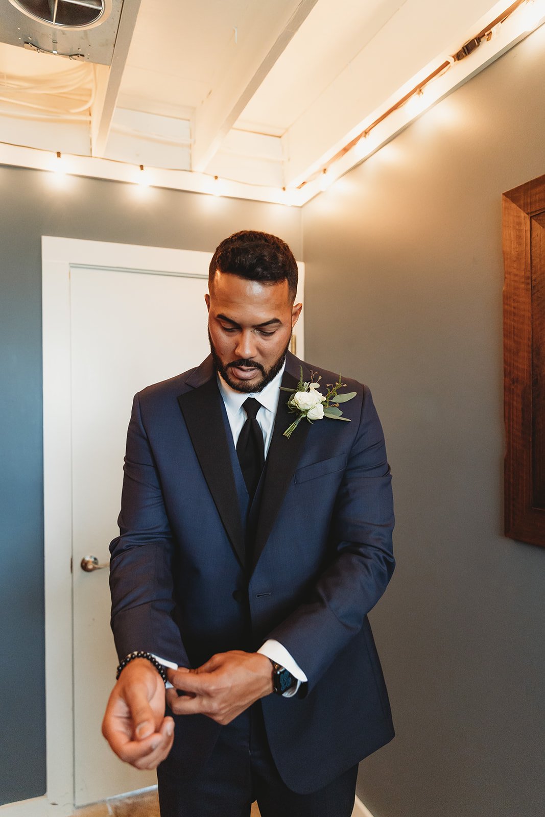 A man dressed in a navy suit with a white shirt and black tie, adjusting his sleeve. He has a small white flower boutonniere on his lapel. The setting appears to be indoors, with a gray wall, white ceiling, and string lights.