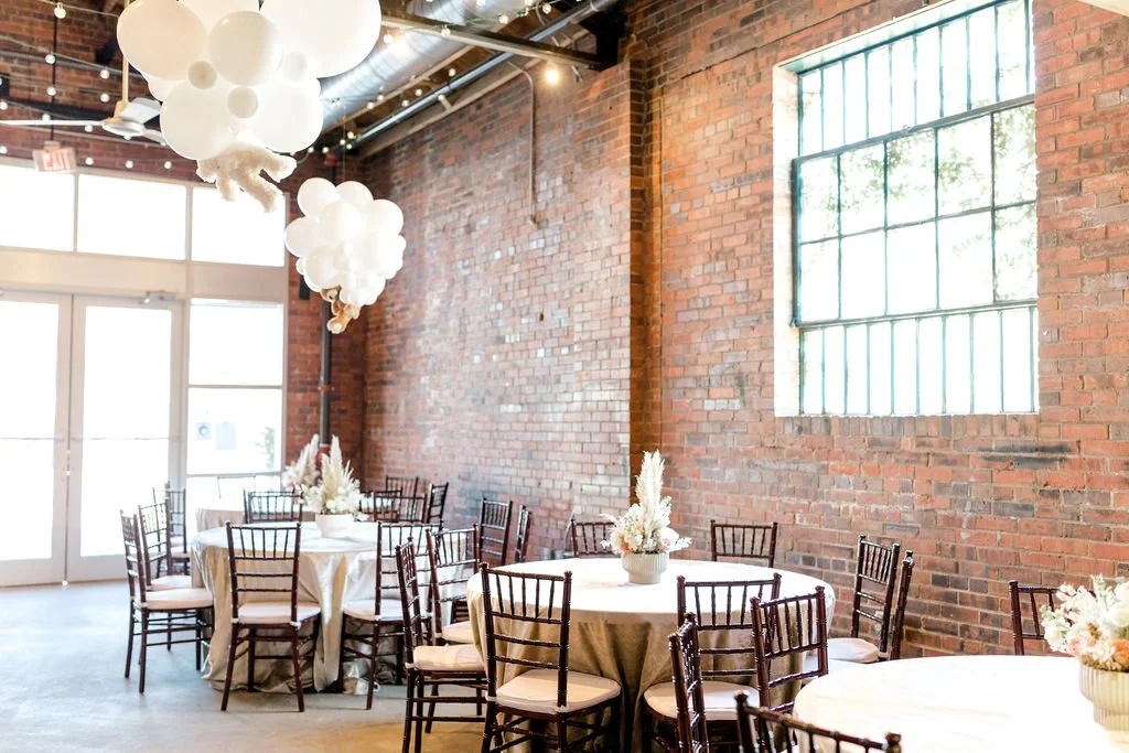 Event space with round tables covered in white tablecloths, floral centerpieces, and wooden chairs, brick walls, large window with metal grid, and balloon arrangements hanging from the ceiling.