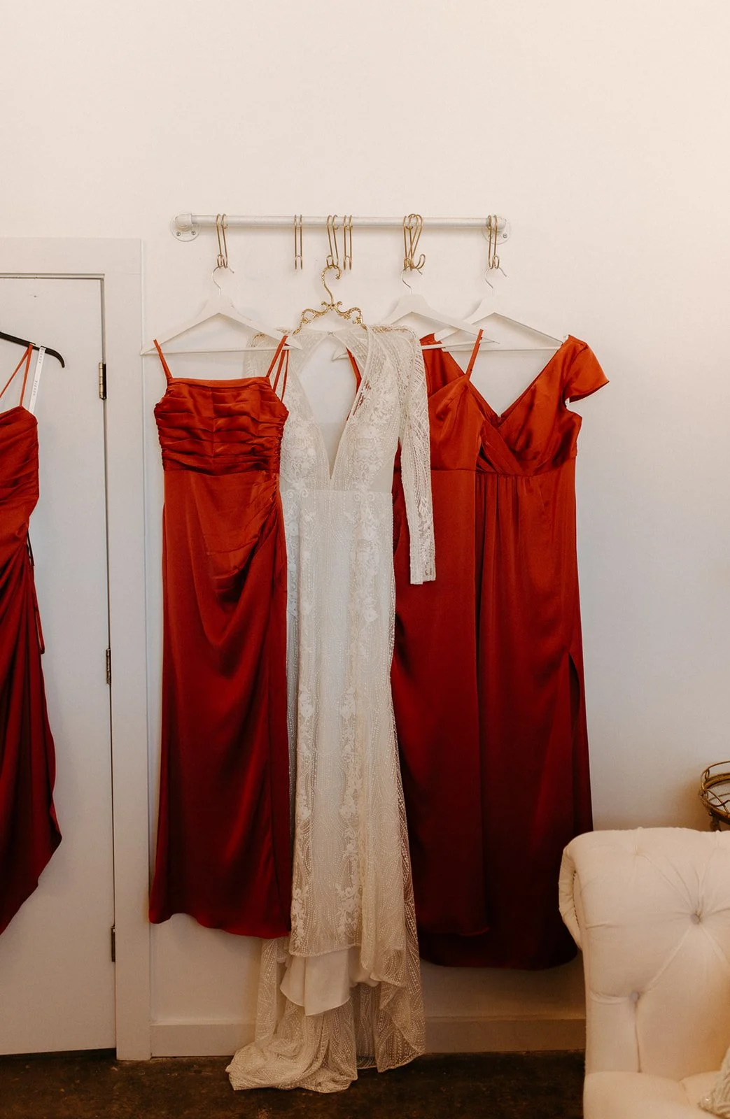 Several red and white dresses hanging on a rack against a plain wall, with a partly visible door and a cream-colored chair nearby.