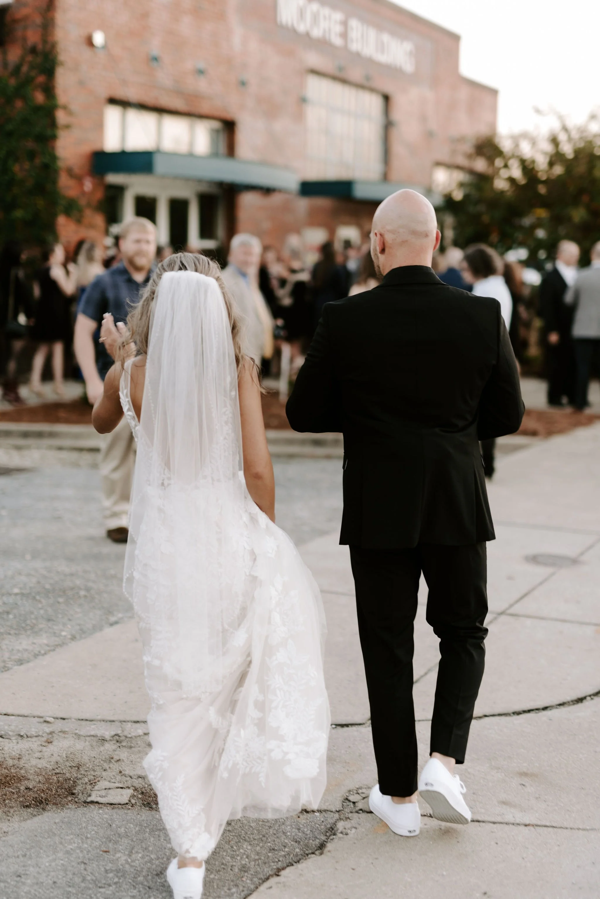 A bride and groom walking together outside a brick building with a crowd of people in the background during a wedding celebration.